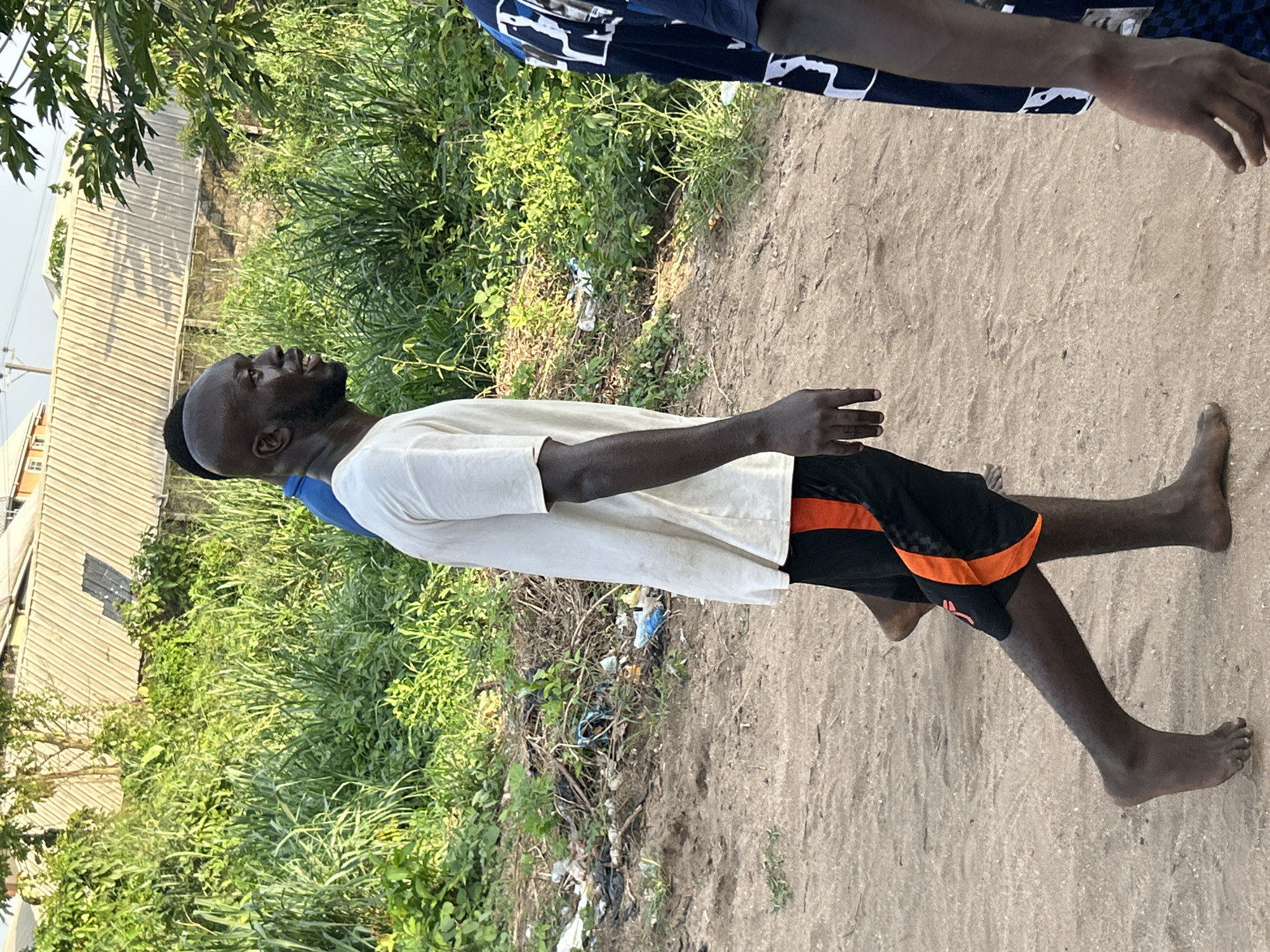 An image shows a Black man standing barefoot on sandy soil. He wears a white t-shirt and black shorts with orange trim. He is looking upwards, possibly interacting with another person partially visible in the upper right. The environment includes lush green vegetation and what appears to be scattered trash along the edge of the sandy area. Buildings are visible in the background on the left, suggesting a residential area of Ihiagwa, Nigeria.