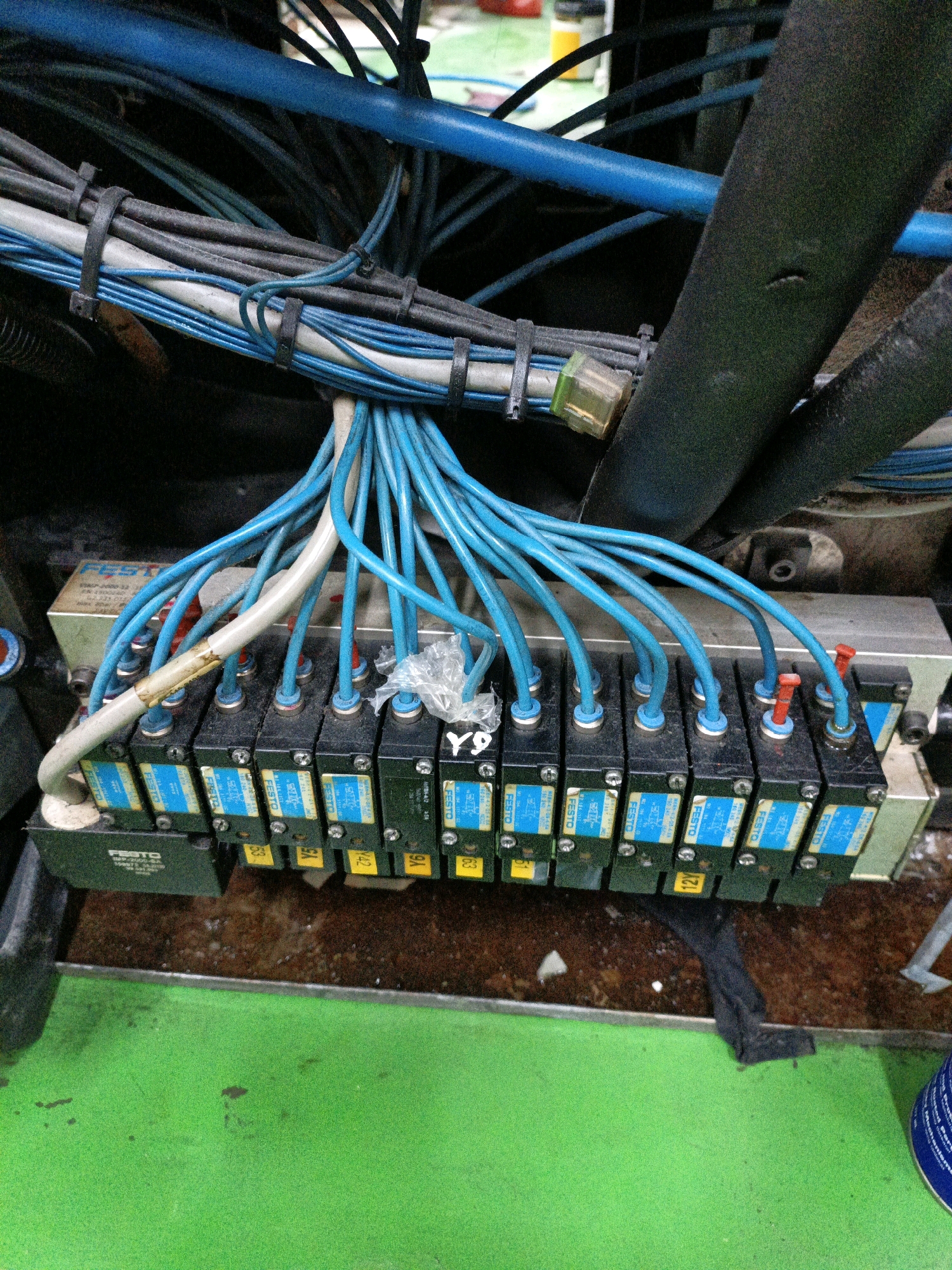 The image depicts a close-up view of industrial pneumatic components, specifically a Festo solenoid valve manifold. The manifold consists of multiple black solenoid valves, each with blue tubing connected to them. These tubes are bundled together and run upwards and to the left, disappearing into a mass of other wires and blue conduits. Some of the valves have yellow labels with numbers or letters, such as "Y9," "Y42," and "Y63." There are also red caps on some of the valve outlets. The manifold is mounted on a silver-colored base, and above it, a large black, ribbed hose is visible, along with more blue tubing and black electrical conduits secured with zip ties. The overall environment appears to be an industrial setting, possibly a factory floor, with a bright green surface in the foreground, suggesting machinery or a painted floor. There are no people or animals in the image. The image is taken from a slightly elevated angle looking down at the equipment. The specific location in the city of North Jakarta, Indonesia cannot be determined from the visual information provided in the image.