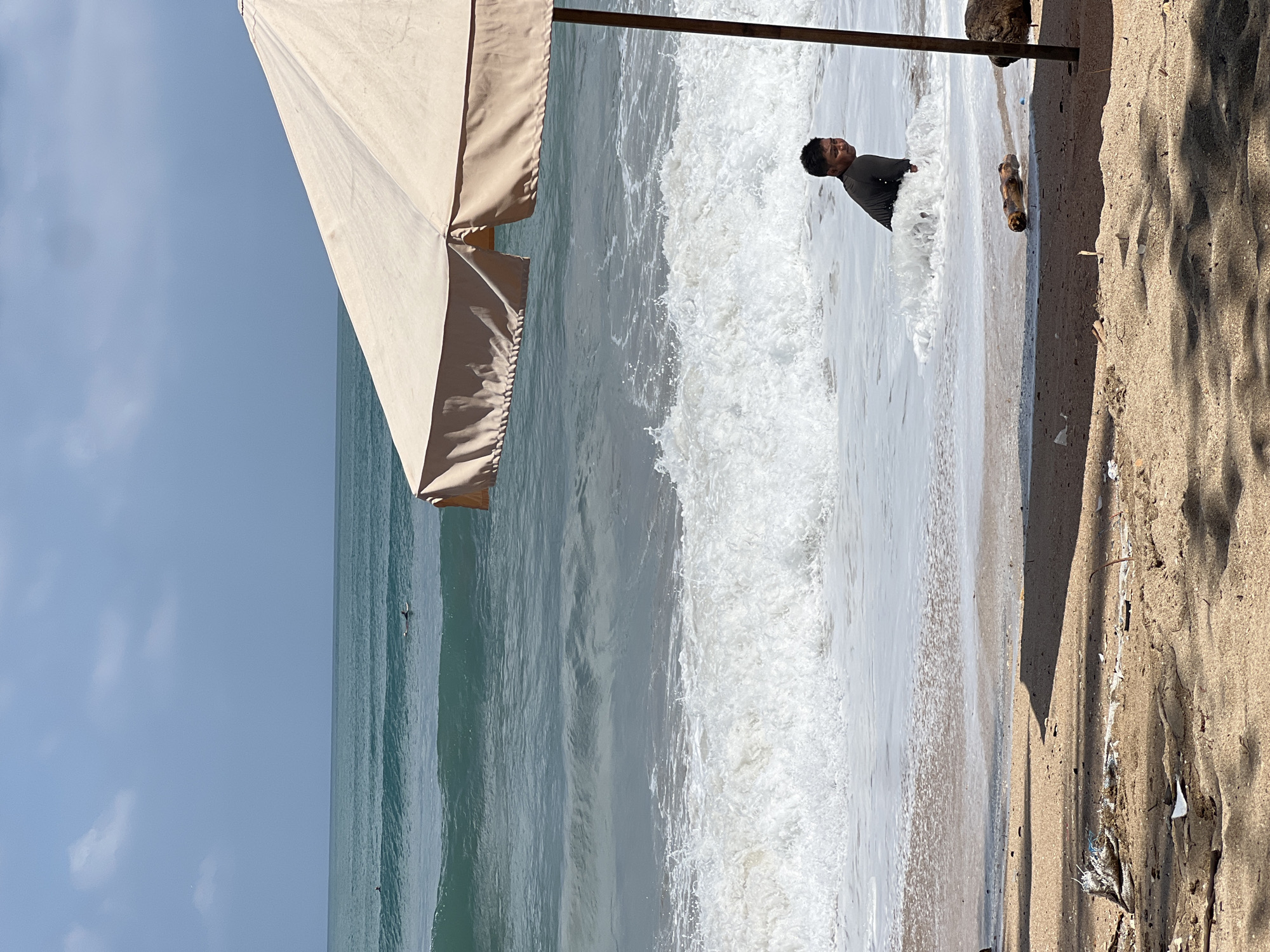 In Kuta, Indonesia, a young person in a dark shirt is partially submerged in foamy ocean waves on a sandy beach. They are looking to their right, with a large, beige beach umbrella casting a shadow on the sand in the foreground. The ocean has significant waves, and there are several surfers visible in the distance. The beach itself is strewn with some debris. The sky is overcast with a pale blue hue.