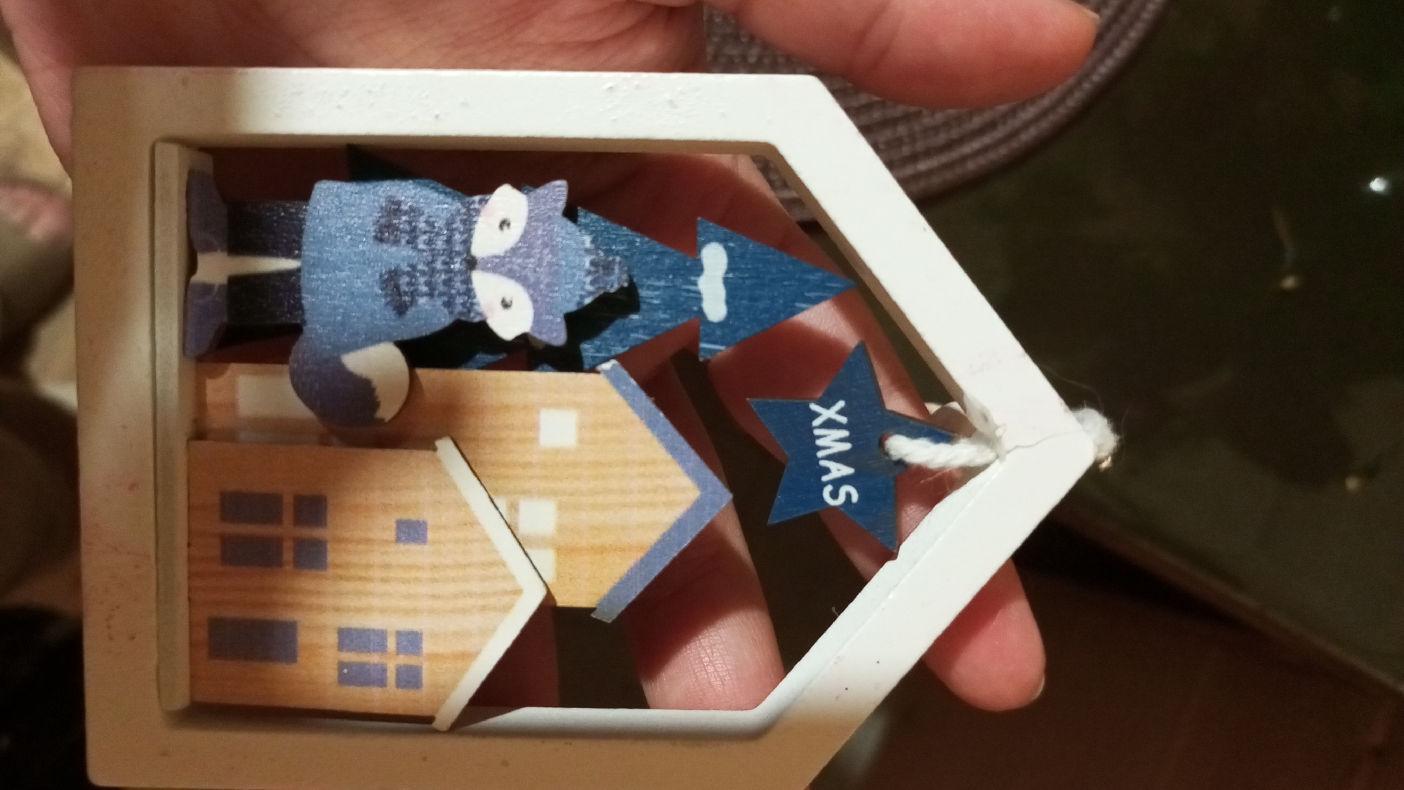 A hand holds a Christmas ornament shaped like a white house frame. Inside the frame are wooden cutouts of two houses with blue accents, a blue fox with white details, and three blue triangular trees, one with a white circle and the other with a blue star with the word "XMAS". The ornament is held against a blurred background of brown and black tones. The location is specified as Юдино, Russia.