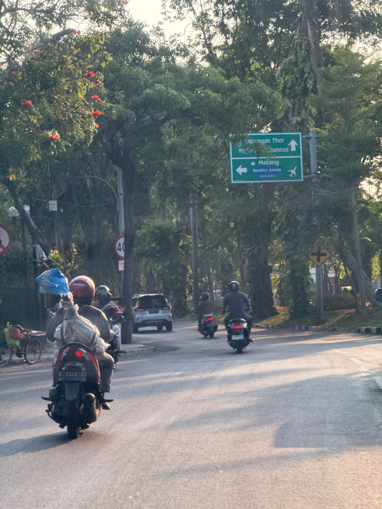 A street scene in Surabaya, Indonesia, captures the late afternoon light filtering through trees lining the road. A directional sign indicates "Lapangan Thor," "Majapahit Muhammad," and "Malang Bandara Juanda (Juanda Airport)," suggesting a route towards these locations. Several motorcycles are visible, with riders and passengers, some carrying bags or objects. One motorcycle in the foreground has a license plate that appears to read "L 2200 BB". A silver car is also on the road. A speed limit sign reads "40". The environment is verdant, with lush trees and some flowering plants. The overall impression is of everyday urban traffic and movement in a well-treed city setting.