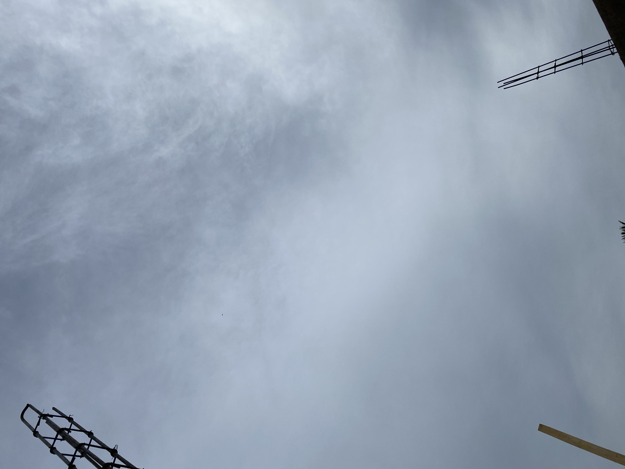 The image shows a view looking up at a cloudy sky with construction rebar visible in the top left and bottom right corners. A wooden beam is also visible in the bottom left corner. The sky is overcast, with some thin, wispy clouds. The overall impression is one of construction activity taking place on an overcast day. The location is likely Banda Aceh, Indonesia, given the context provided. There are no people or animals visible in the image, and no specific actions or interactions are depicted.