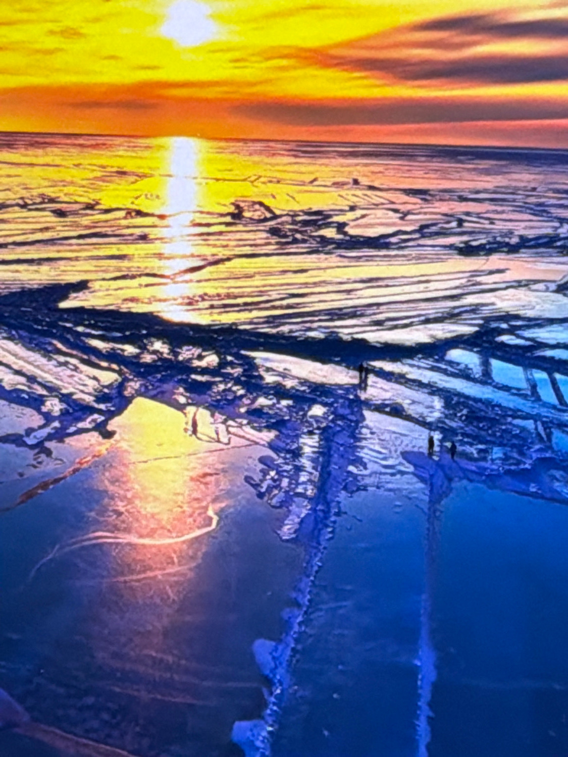 The image depicts a dramatic sunset over a vast, frozen landscape, likely a lake or sea. The sky is ablaze with vibrant hues of yellow, orange, and red, with the sun casting a bright, elongated reflection across the icy surface. The ice itself is fractured and uneven, with dark shadows and lighter patches creating a textured appearance. Several small figures, appearing as silhouetted individuals, are scattered across the ice, observing the sunset. The scene evokes a sense of awe and perhaps isolation in a stark, natural environment. The prompt mentions Kafr Yasif, Israel, however, the visual elements of the image do not provide any specific clues to confirm or deny this location. The image is a generic depiction of a frozen sunset.