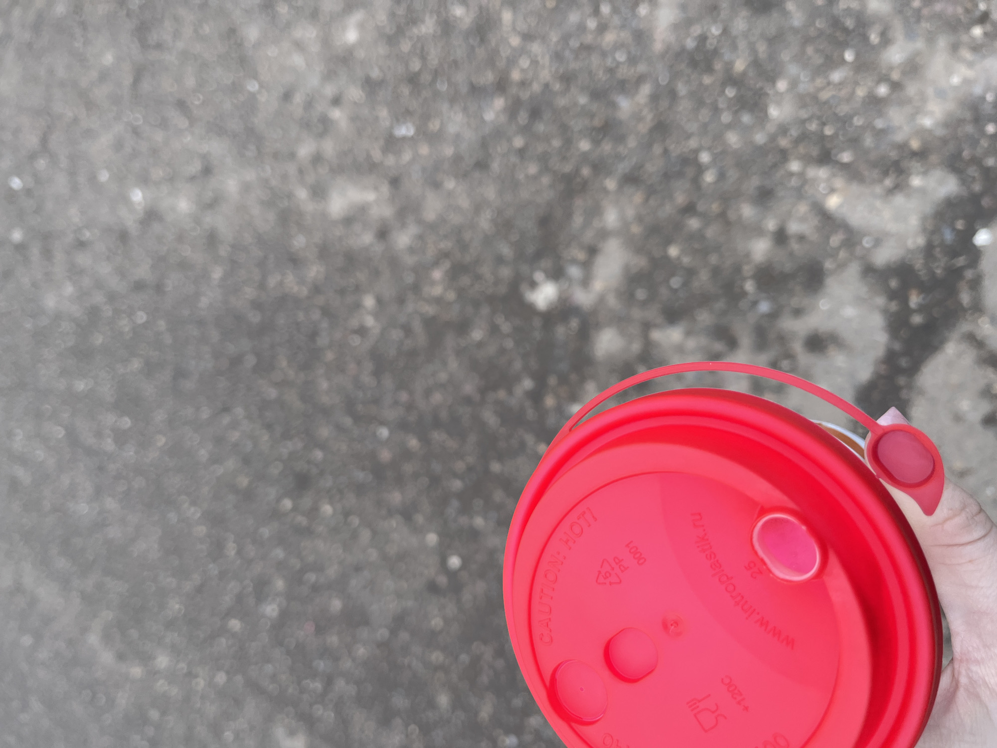 The image depicts a close-up view of a red plastic lid, likely from a disposable cup, held by a hand with a pink nail. The background is a blurred gray surface, possibly a sidewalk or pavement. The lid has markings including "CAUTION: HOT!", "PP 5", and a website address "www.intropolastik.ru". The website address suggests the lid could be manufactured in or distributed from Russia. Given this and the information available it is possible this photograph was taken in Russia. The city Chaikovsky, Russia is unknown, however, is possible this photo was taken there.