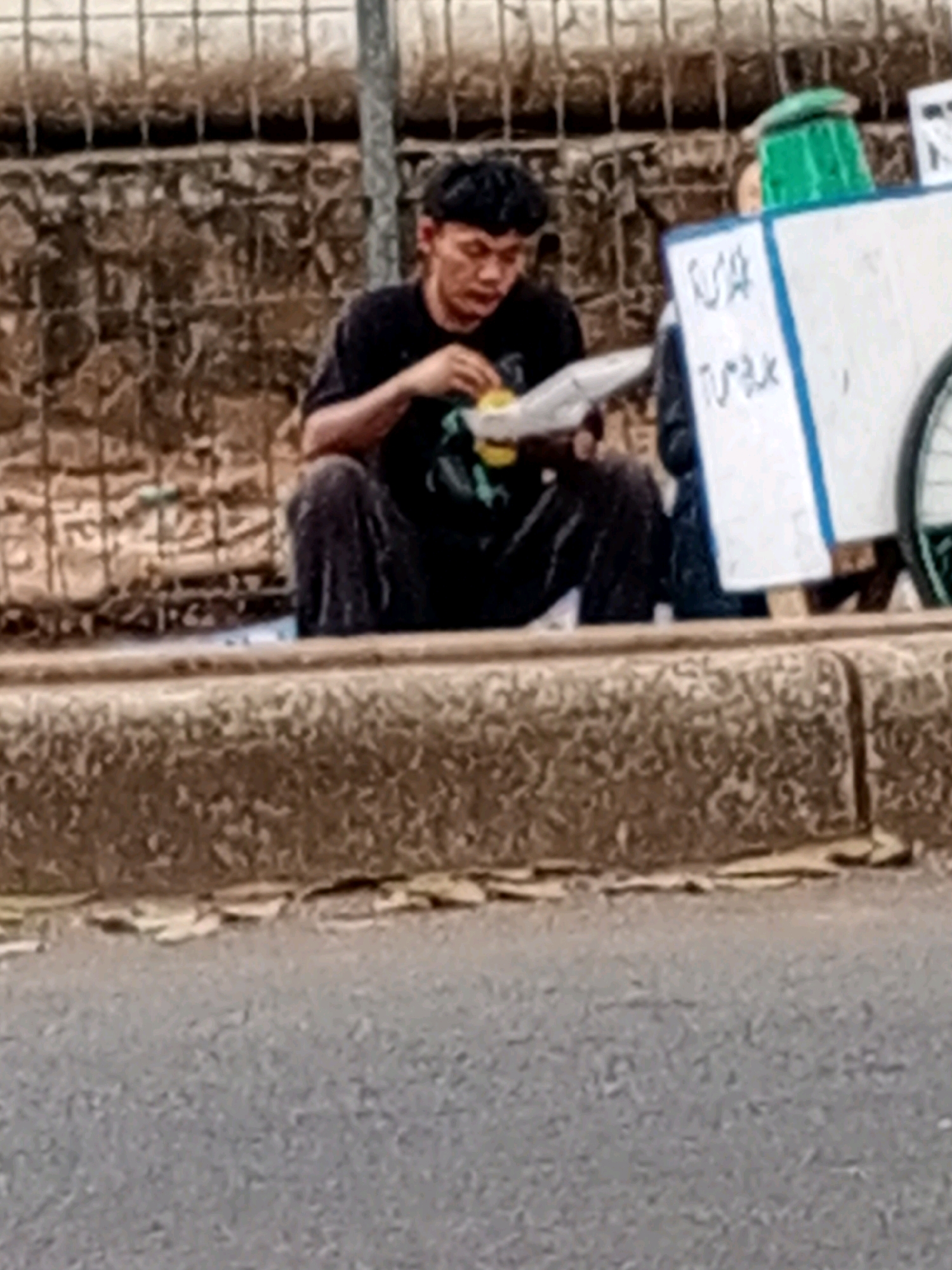 In the city of South Tangerang, Indonesia, a young man with dark hair and wearing a black t-shirt and dark pants is sitting on a curb beside a road. He is holding a white paper container and appears to be eating something from it with his hands. To his right, there is a white cart with blue trim, topped with a green object that resembles a broom or a large brush. A sign on the cart has writing on it, with the words "USA" and "TUMUR" visible. The background consists of a stone wall and a metal fence. The road in the foreground is paved with asphalt.