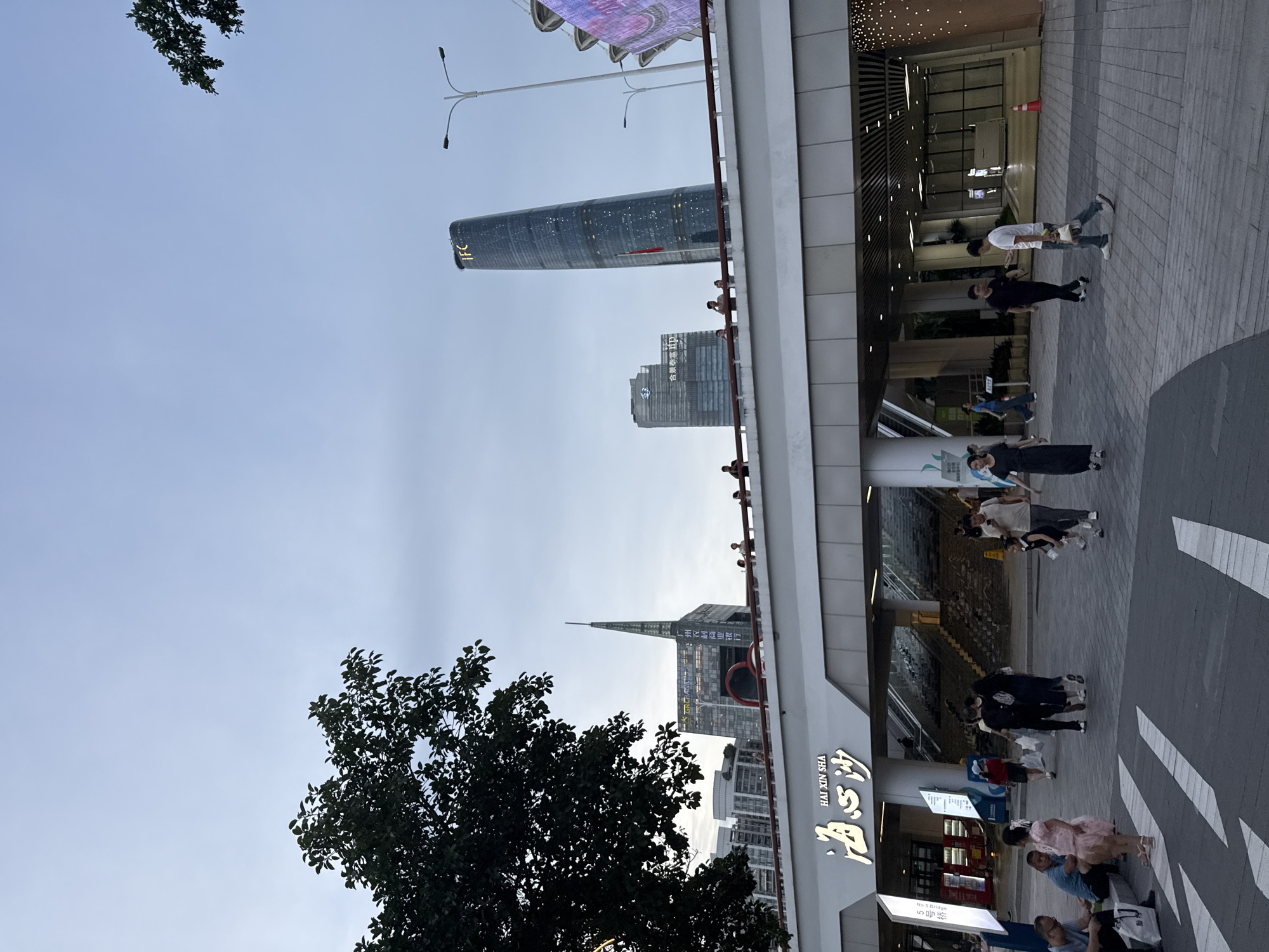 In Tianhe District, China, the image depicts a modern urban scene with a focus on a pedestrian overpass. The overpass is adorned with the Chinese characters "海心沙" (Hǎixīnshā), which translate to "Heart Island." Several people are visible on and around the overpass, some walking, others standing and observing. Behind the overpass, a tall, slender skyscraper with the illuminated letters "IFC" stands prominently against a pale sky. To its left, another skyscraper features a more angular design and signage. Trees are present in the foreground and background, with one large tree canopy visible in the upper left quadrant of the image. The overall environment suggests a bustling commercial or public space in a city.
