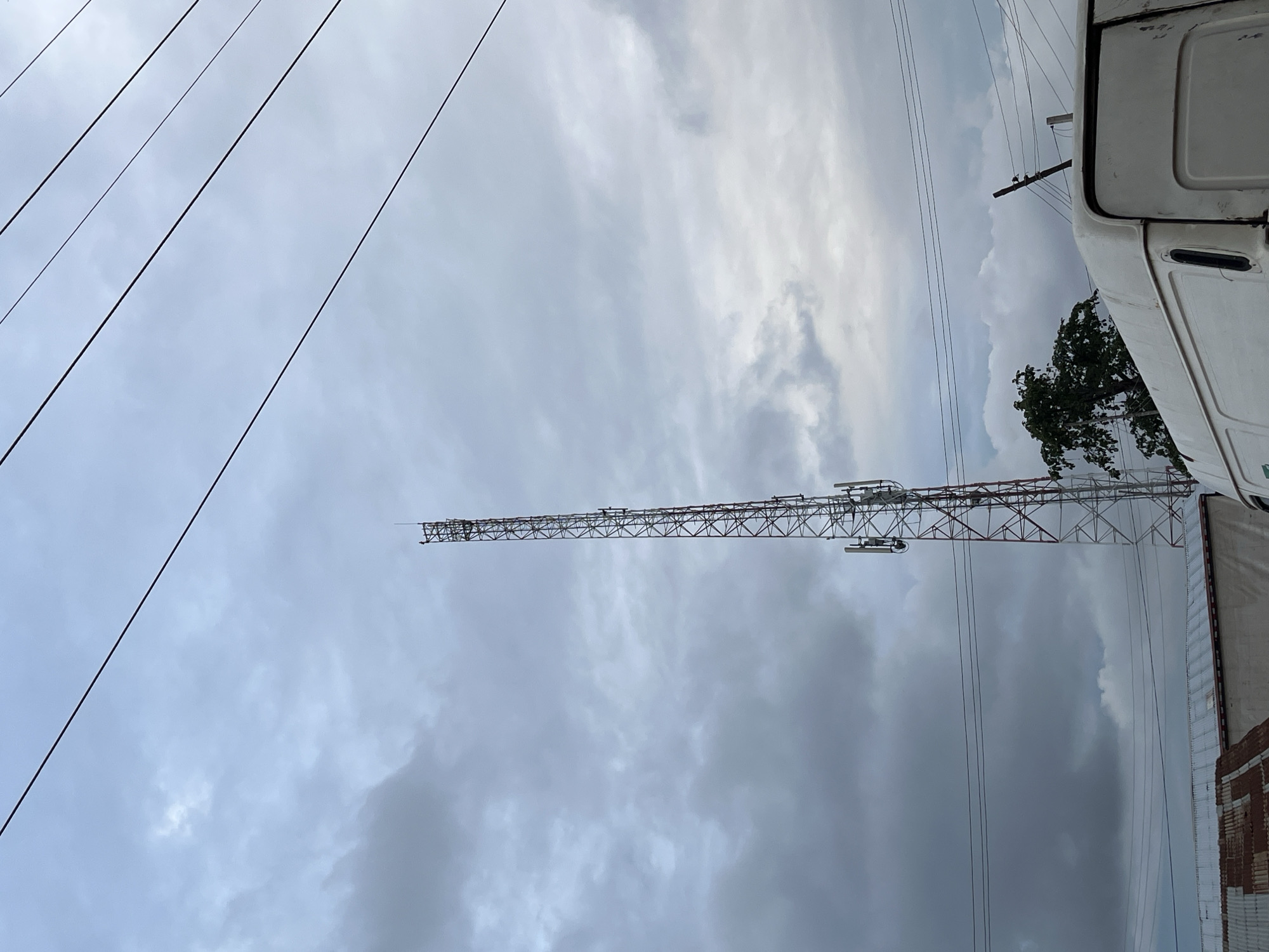 A tall, red and white lattice telecommunications tower stands prominently against a cloudy sky in Bukuru, Nigeria. Multiple sets of antennas are visible on the tower. Several power lines stretch across the upper portion of the image. In the foreground, the top of a white van is visible, along with a section of a corrugated metal roof of a building. A lush green tree is situated behind the van and to the left of the tower. The overall scene depicts an urban or semi-urban environment under an overcast sky.