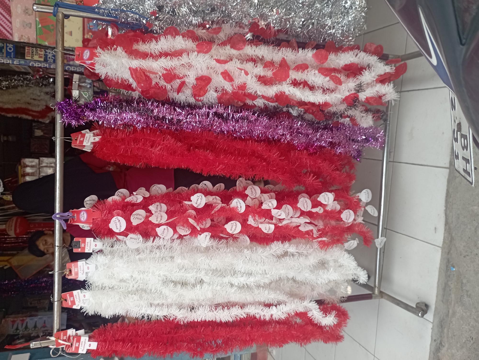 A rack of holiday tinsel is displayed outside a shop in Purwakarta, Indonesia. The tinsel is in various colors, including red, white, purple, and silver, and some strands are adorned with red heart-shaped cutouts. Behind the tinsel, a person wearing a headscarf and a dark garment is visible, along with parts of shop merchandise. The ground in front of the display consists of white tiles and a concrete walkway. A motorcycle license plate with the characters "BH" is visible in the lower right corner. The overall scene suggests a festive outdoor market or street vendor setup.