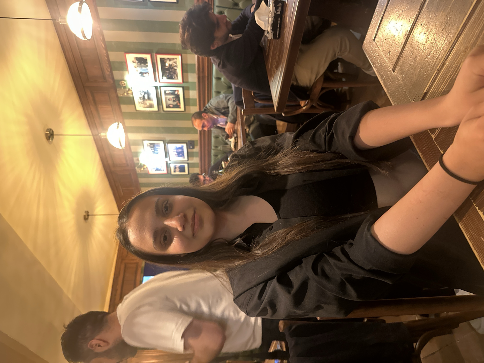The image captures a moment inside a Yerevan, Armenia establishment, likely a restaurant or pub, characterized by its striped wallpaper, framed photos on the walls, and warm lighting from sconces. A young woman with long dark hair is seated at a wooden table, wearing a black blazer over a black top. Her arms rest on the table, and she appears to be looking at the camera. A man in a white shirt is partially visible in the lower left corner, appearing to be seated next to her. In the background, other patrons are seated at tables, including a man in a dark jacket examining something on the table, and other figures further back engaged in conversation. The overall atmosphere suggests a casual, social setting.