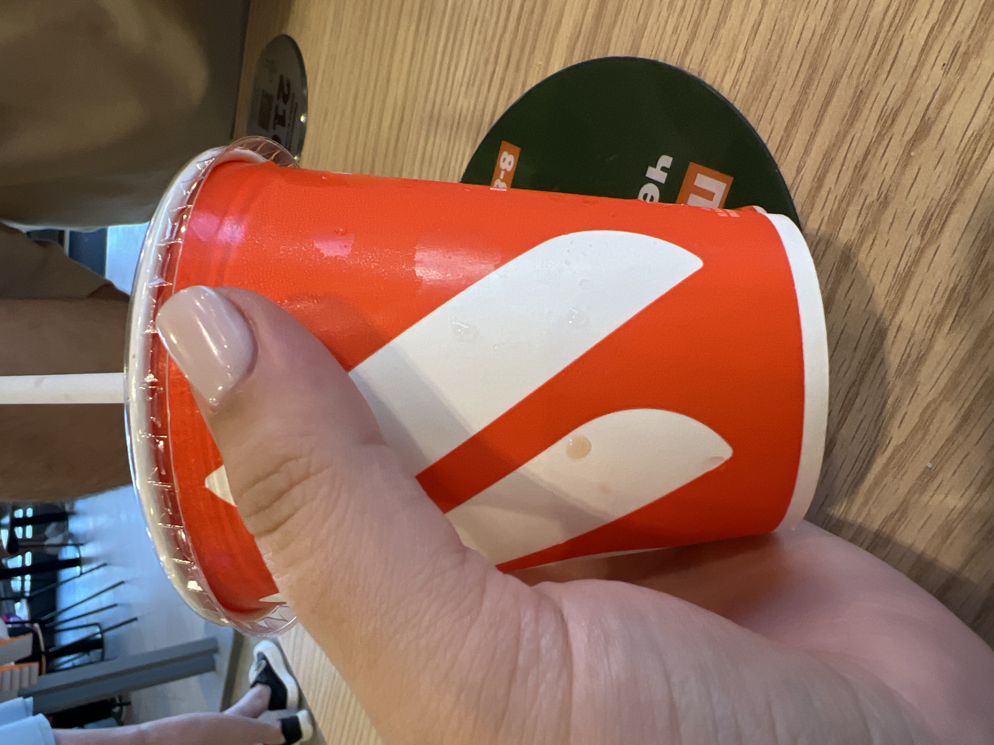 A hand with light pink nail polish is holding a red and white paper cup with a clear plastic lid and a white straw. The cup has condensation on its exterior. The cup is placed on a wooden surface, possibly a table, with a dark green circular mat partially visible underneath it. The mat has white text and symbols on it, including what appears to be "8-8" and "yeh" and an orange square with a white "n". In the background, out of focus, there are chairs and people, suggesting an indoor public space like a cafe or restaurant. The lighting is bright.