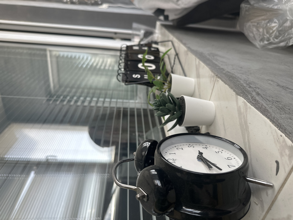 The scene depicts an interior room, specifically a windowsill or narrow shelf located next to a window fitted with horizontal venetian blinds, in the city of Unknown, Unknown. In the left foreground, a black twin-bell alarm clock with silver legs and a silver handle is positioned, displaying white numbers and black hands, indicating approximately 1:50 or 2:50. To the right of the clock are two small potted plants in white cylindrical pots; the first is a green succulent, and the second, further back, is another green leafy plant. Beyond these plants, a black-framed flip-calendar or similar device displays white characters against black tiles, including an "S" and a "0" or "U". The surface holding these items is light-colored and appears tiled or patterned. A blurry reflection of a human head or upper torso is visible within the window's glass and blinds in the background, facing away from the camera or in profile. The wall directly beneath the windowsill is textured and light gray. On the extreme right, an out-of-focus white object, possibly a plastic-wrapped item or bag, is partially visible.