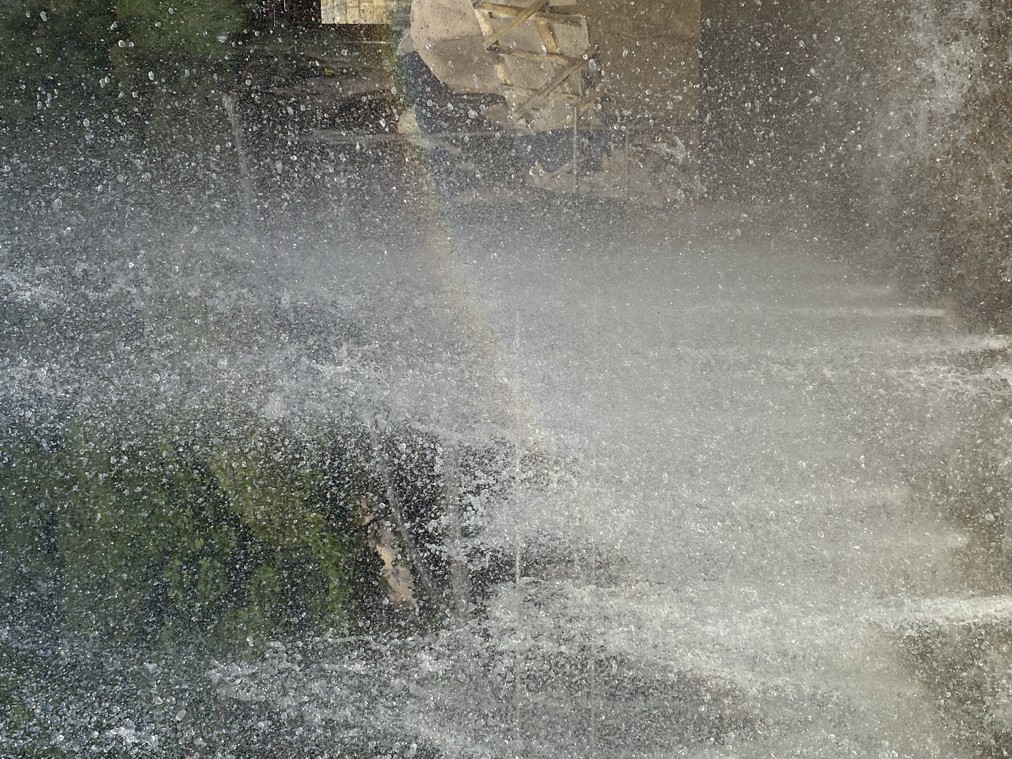 The image shows a fountain with water spraying in the air, creating a mist. Behind the water, there are green trees and some man-made structures. The lighting suggests it is daytime. The foreground is dominated by the water spray. In the background, to the right of the frame, there are stone structures and some wooden elements. A faint rainbow is visible through the water spray. There are no people visible in the image. The environment appears to be outdoors, possibly in a park or public space.