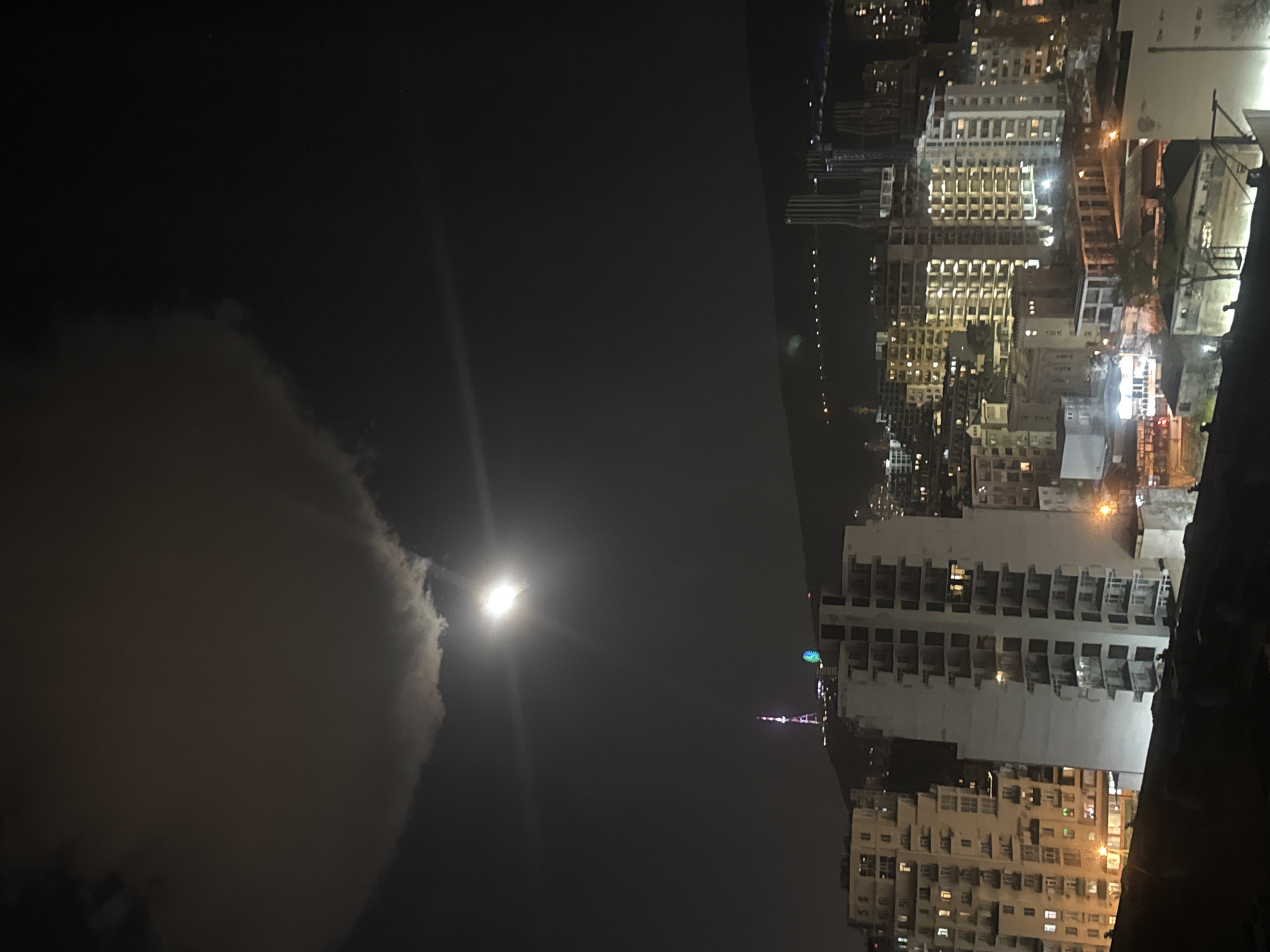 The image shows a nighttime view of Tbilisi, Georgia, with a bright moon partially obscured by clouds. Buildings with lit windows dominate the right side of the frame, indicating a densely populated urban area. A tower with colorful lights is visible in the distance. The sky is dark, with the moon serving as a prominent light source.