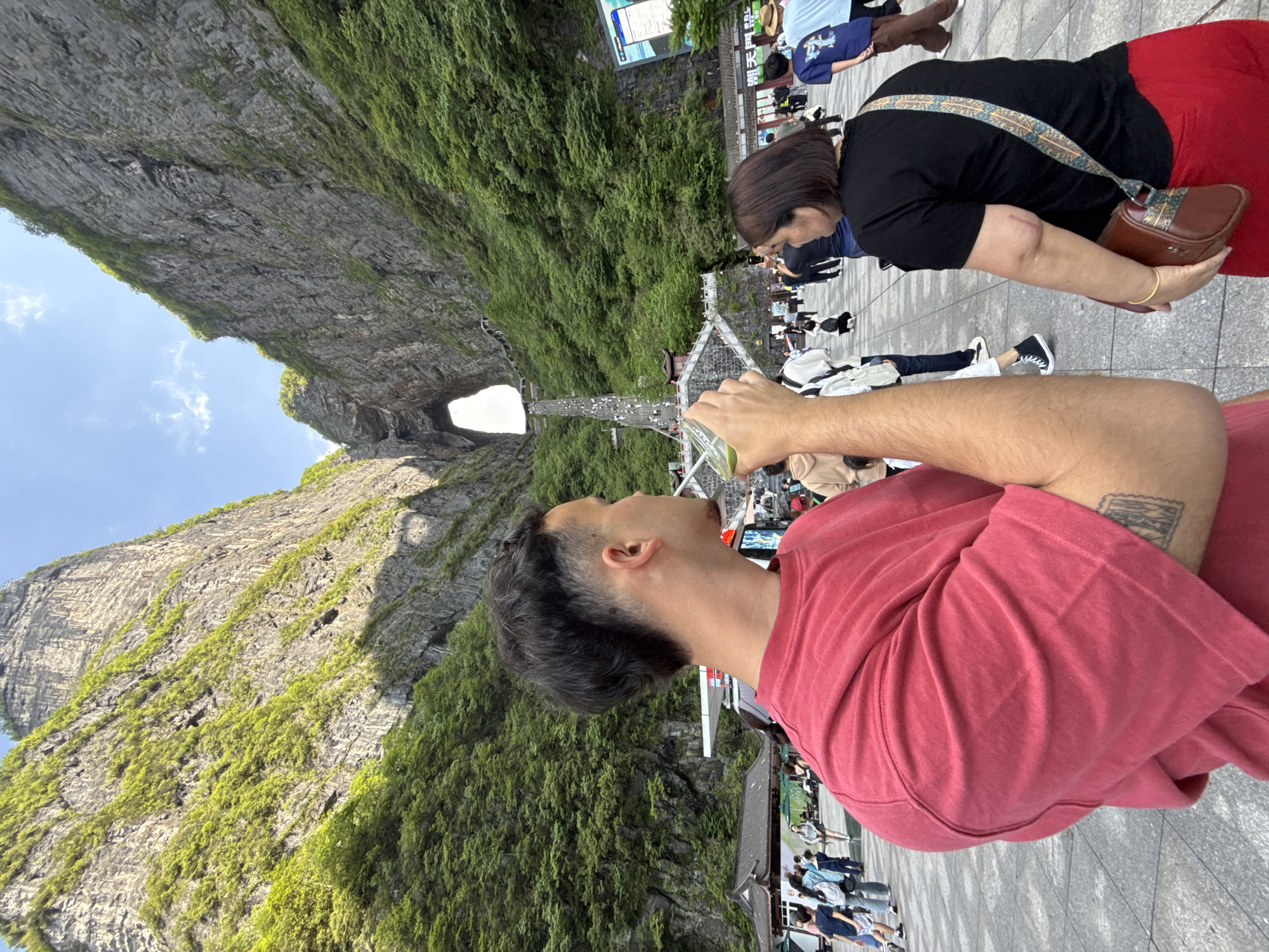 The image depicts the Tianmen Cave, a large natural arch in Tianmen Mountain National Park, in Zhangjiajie, China. The cave is visible through a gap in the rock formation on the left side of the image. A man wearing a red shirt is in the foreground looking in the direction of the cave. Other tourists are visible near the man and in the background. The sky is visible above the cave.