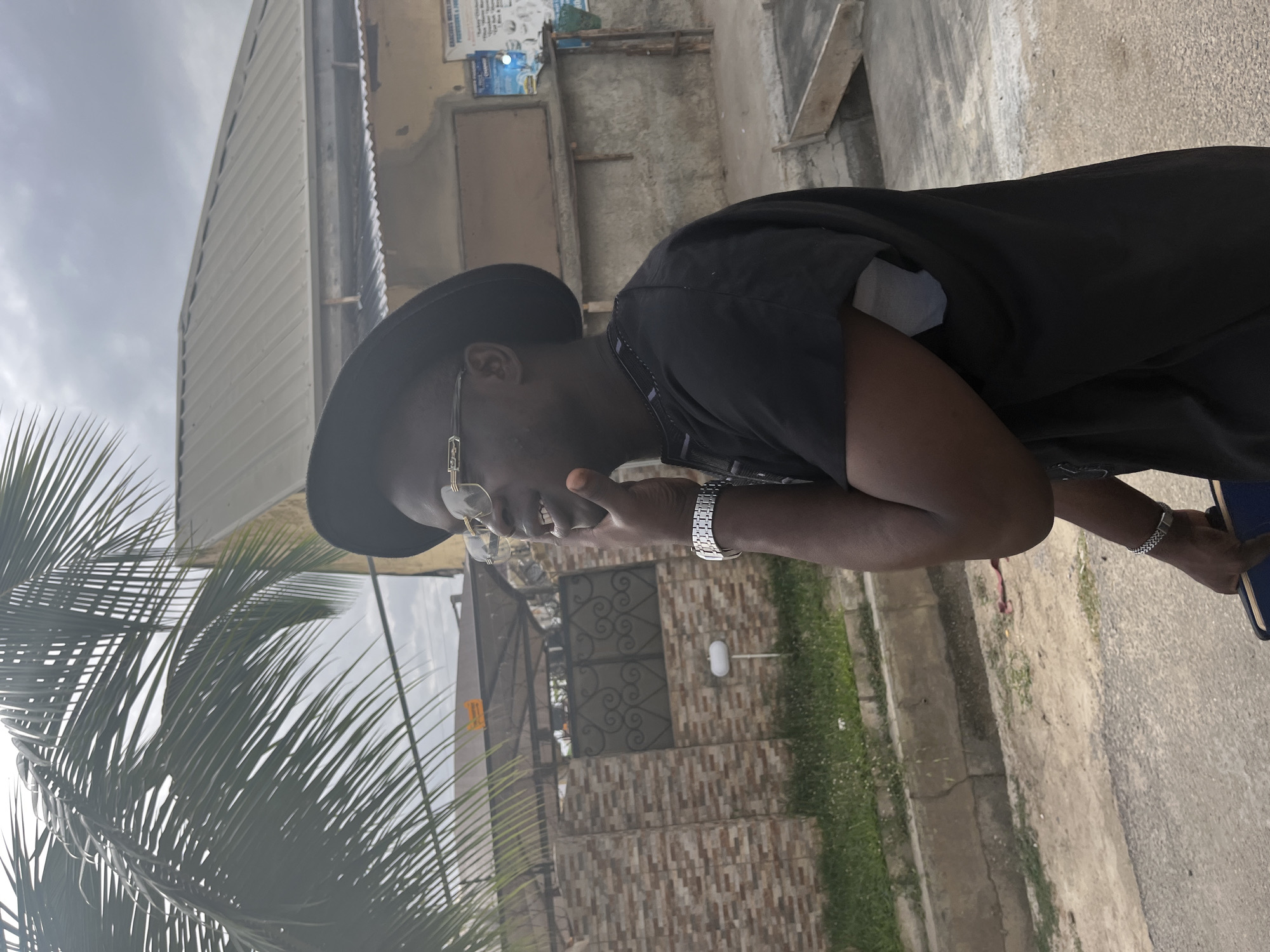 A man wearing a black hat and black clothing stands on a paved surface next to a street. He wears glasses and a metal watch, and is touching his face. In the background, there is a building with advertisements, another building with a corrugated metal roof, a gate, and various plants. Palm fronds are visible in the foreground. The location appears to be an urban or suburban area, and is potentially located in Abuja, Nigeria.