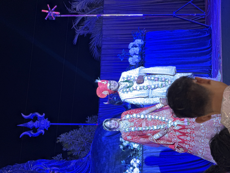The image captures a vibrant night-time event, likely a wedding reception or ceremony, set against a dark background illuminated by blue lighting. The central focus is a couple dressed in traditional Indian wedding attire, positioned on a decorated stage.
The groom, on the left, is wearing a richly embroidered white or cream sherwani, a prominent red turban (pagdi), and glasses. He is adorned with multiple floral garlands, including one with green beads and another with white and pink flowers. His hands show intricate mehndi designs. He is looking towards the front with a slight smile.
The bride, positioned to the groom's right, is dressed in a heavily embroidered red lehenga with gold or silver detailing. She wears a matching dupatta and traditional jewelry, including a necklace, earrings, bangles, and a maang tikka on her forehead. Like the groom, she is wearing floral garlands made of white and pink roses and has mehndi on her hands. She is also looking forward with a smile.
The stage is adorned with draped blue fabric, creating a rich backdrop. White floral arrangements are visible on the stage, particularly behind and around the couple. Dark palm fronds or artificial foliage are also part of the set design, appearing against the dark background.
Tall, decorative poles flank the couple. To the left, a blue, trident-like structure (resembling a Trishul) sits atop a pole. To the right, another pole is topped with a luminous, star-shaped or snowflake-like fixture that emits light. Blue spotlights crisscross the scene, casting rays of light and contributing to the dramatic atmosphere.
In the foreground, partially obscuring the lower right portion of the bride, is the back of a person's head and shoulder, suggesting an observer or guest. The overall ambiance is festive and celebratory, characteristic of a wedding. No visible text is present in the image.