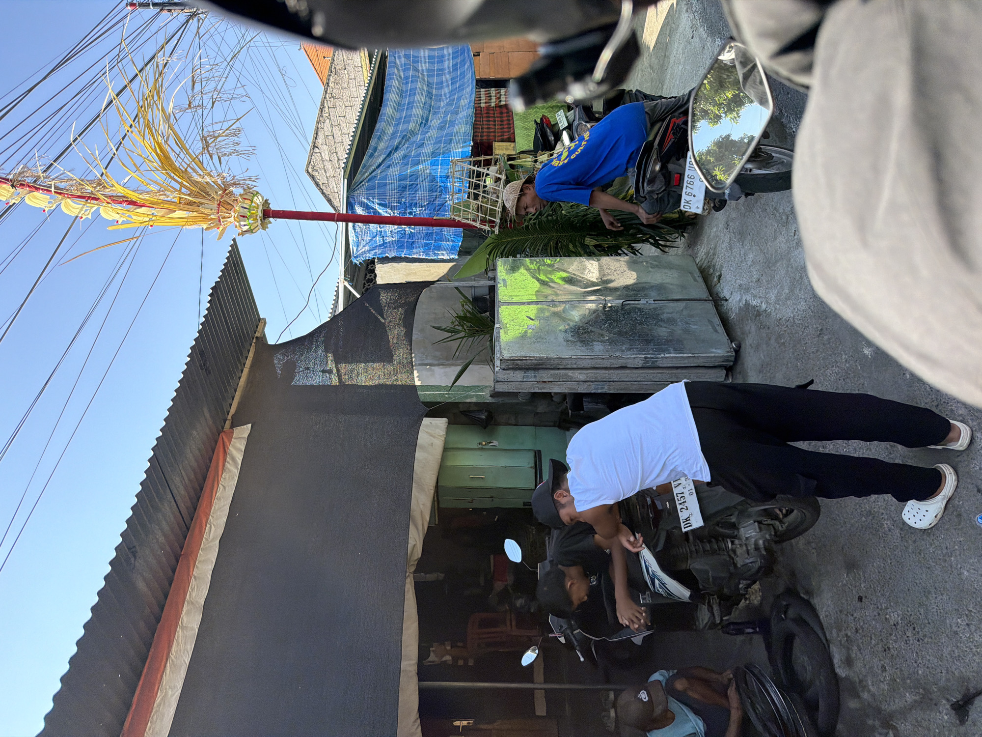 In Sawan, Indonesia, on a sunny day, a group of individuals are gathered near a roadside workshop. The scene depicts a motorcycle repair or service area. Two men are leaning over a motorcycle, seemingly engaged in its maintenance. One man wears a white t-shirt and black pants, while another wears a black t-shirt and a black cap. A younger individual in a blue shirt and a cap is crouched nearby, looking at a tire. Further down the street, another man in a blue t-shirt and a straw hat is sitting on a motorcycle, looking towards the left of the frame.
The workshop itself appears to be open-air, with a corrugated metal roof extending over a dark-colored wall. Various items are visible, including metal cabinets, potted plants, and stacked materials. Overhead, a tall, decorative bamboo structure is adorned with yellow leaves and red fabric, possibly a ceremonial decoration. Electrical wires crisscross the sky above. The ground is paved with concrete, and the overall atmosphere suggests a typical day in a local Indonesian community. The license plates on the motorcycles are visible, indicating they are registered in Bali, Indonesia.