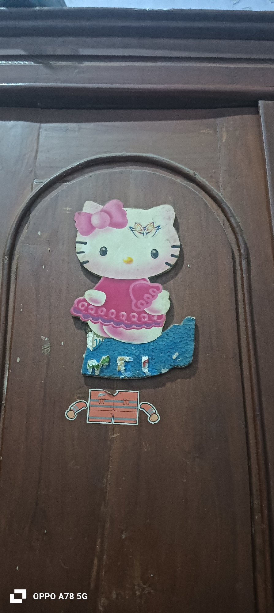 In Buncitan, Indonesia, a Hello Kitty sticker is affixed to a wooden door. The sticker depicts Hello Kitty wearing a pink bow and dress, with a blue textured material below her. Below Hello Kitty, a separate sticker shows a pair of orange shorts with two arm attachments. The door exhibits a dark wood grain texture.