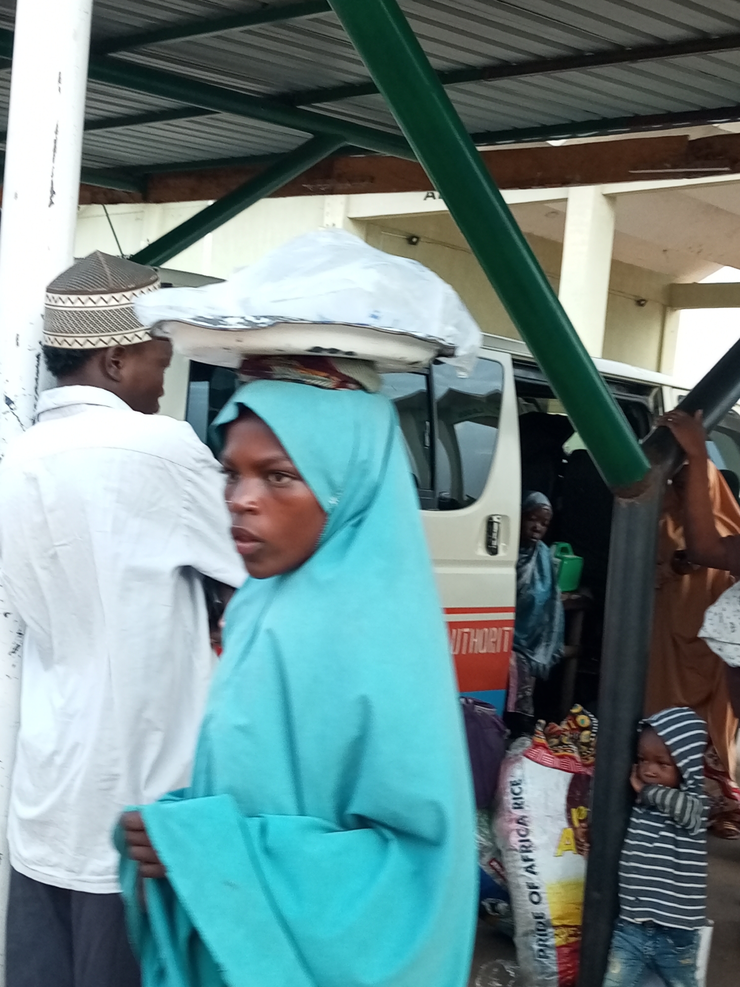 In Gombe, Nigeria, a group of people are gathered under a covered structure, possibly a bus station or market. A woman in a turquoise hijab is carrying a large white package on her head, with a man wearing a patterned cap beside her. A white minivan is parked behind them, with its side door open. To the right, a young child in a striped hoodie stands by a pillar, leaning their head against their hand. Bags of rice are visible near the child, one labeled "PRIDE OF AFRICA RICE". The environment suggests a bustling public space with an overhead metal roof supported by beams and pillars. The lighting is somewhat diffused, indicating an overcast day or a shaded area.