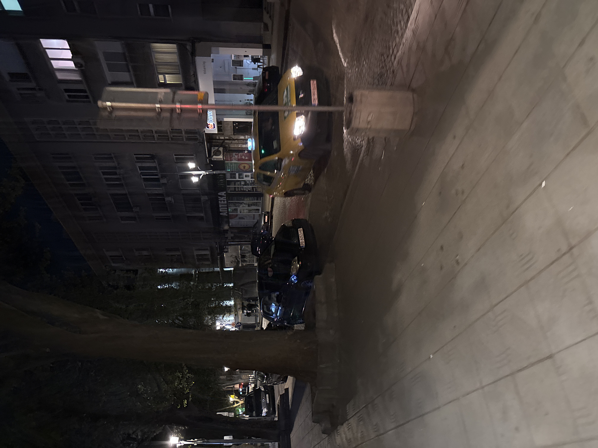 The image depicts a street scene at night in Plovdiv, Bulgaria. A yellow taxi is stationary on the street, headlights illuminating the scene. A black car is parked further down the street. Buildings line the left side of the street. A tree is visible along the sidewalk. Pedestrians may be present. A sign is visible on one of the buildings, with the word "Аптека". The street appears wet, possibly from rain.