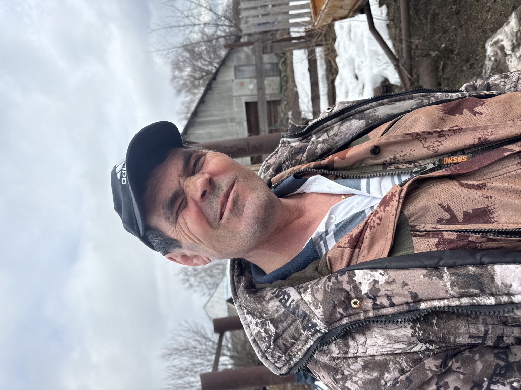 A middle-aged man wearing a camouflage jacket and a black baseball cap is visible in the foreground. He is facing the camera. Behind him is a rural scene with a wooden fence, a shed or small building, and some patches of snow on the ground. The sky is overcast. The location is assumed to be the city of Кемуль, Russia.