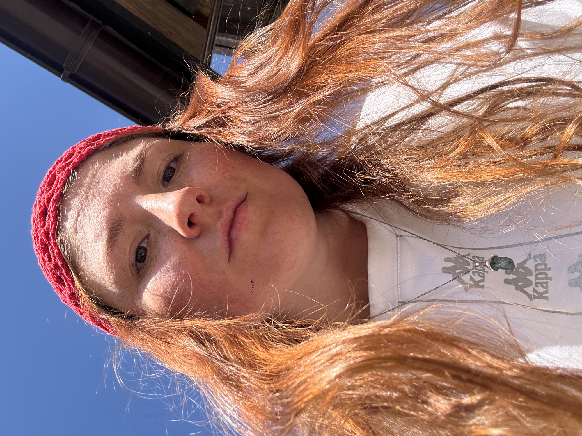 A close-up, low-angle selfie shows a person with long, reddish-orange hair and a red bandana on their head. They are wearing a white t-shirt with a Kappa logo and a necklace with a green pendant. The background is a bright blue sky and a brown overhang. The image appears to be taken outdoors on a sunny day. There is no indication of the location being Nizhny Novgorod, Russia.