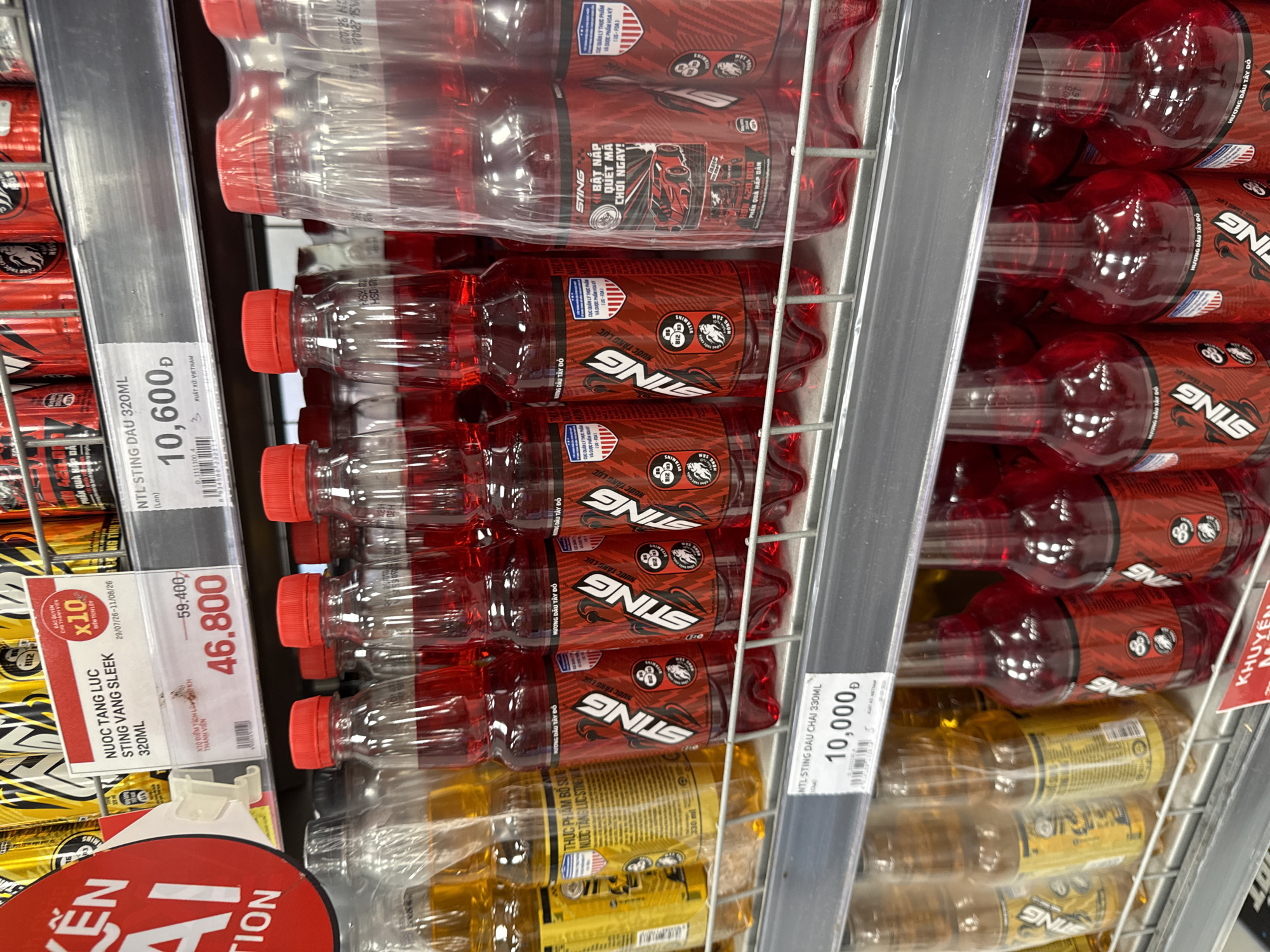 The image is a close-up, eye-level shot of a retail shelf in a brightly lit store, likely a convenience store or supermarket, in Nha Trang, Vietnam. The focus is on an assortment of bottled beverages, primarily energy drinks.
On the top shelf, several rows of canned energy drinks are visible, mostly in red and yellow packaging. A price tag for "NUOC TANG LUC STING VANG SLEEK 320ML" displays a discounted price of "46,800" Dong, with the original price of "59,400" Dong. Another price tag indicates "NTL STING DAU 320ML" at "10,600" Dong.
The middle shelf prominently features multiple bottles of red-colored energy drink labeled "STING" with Vietnamese text and graphics. These bottles are arranged in rows and appear to be sold individually and in multi-packs wrapped in plastic. A smaller price tag for "NTL STING DAU CHAI 330ML" shows a price of "10,000" Dong. To the left of these red drinks are bottles of a yellow-colored beverage, also labeled "STING" with Vietnamese text.
The bottom shelf shows more of the red "STING" energy drinks and some yellow drinks. A red and white sign with Vietnamese text, likely advertising a promotion, is partially visible at the very bottom of the frame.
The overall environment suggests a commercial retail setting with organized product displays. The lighting is even and bright, typical of a store interior. No people are visible in the image. The perspective is static, focusing on the product details and pricing.