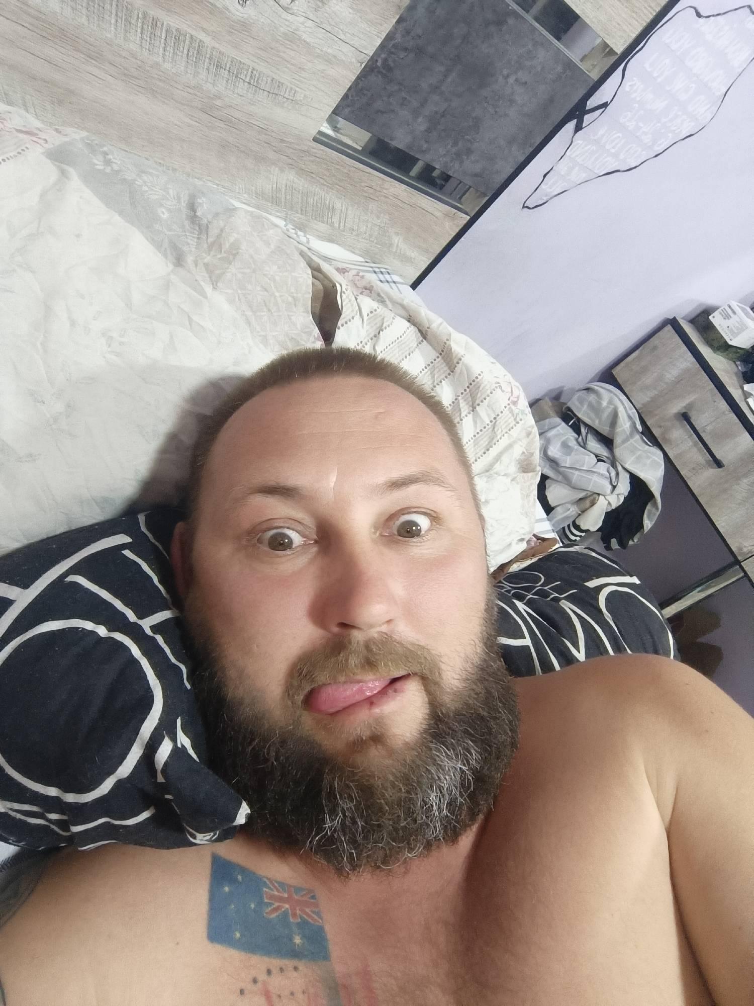 In a bedroom, a man with a beard and a flag tattoo on his chest lies on his back, sticking his tongue out at the camera. The man is wearing a black and white striped pillow. In the background, there is a wooden headboard and a white wall with a drawing of a map and writing on it. A bedside table with clothes is visible to the right. There are no visible indicators to determine the location in Taraz city administration, Kazakhstan.