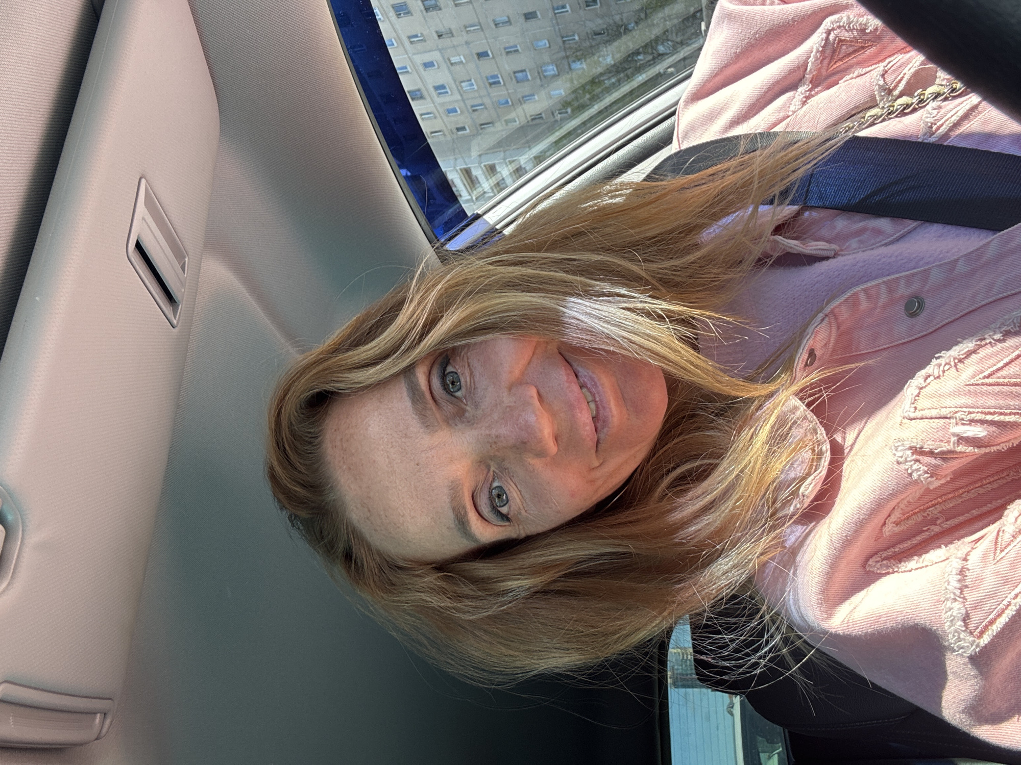 A woman with fair skin, light brown hair, and freckles is inside a car, wearing a pink denim jacket and a seatbelt. Outside the car window is a multi-story building with numerous windows, typical of residential or office buildings in urban settings. Based on the architectural style of the building and the surrounding environment, it is plausible that the location is Saint Petersburg, Russia. The woman is facing the camera with a slight smile. The interior of the car is light gray.