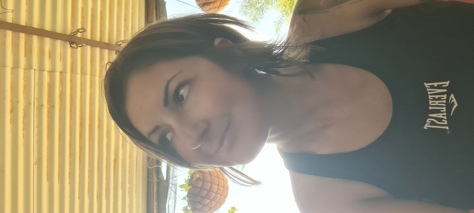 A casual, upright portrait captures a young woman smiling subtly, looking up and slightly to her right. She has dark, medium-length hair and a septum piercing. She is wearing a black tank top with the word "EVERLAST" and its logo printed in a light color on the front (the text appears reversed in the provided image).
The setting appears to be outdoors, likely in bright daylight, indicated by the strong light source behind her creating highlights on her hair and face. The background features a yellow-beige corrugated metal wall on the left, showing signs of weathering or rust. A wooden beam with metal hooks or chains runs horizontally across the upper part of the wall. To her right and behind her, a bright sky is visible through green foliage, suggesting trees or plants, and a woven hanging basket with some greenery is partially seen. The overall mood is relaxed and pleasant.