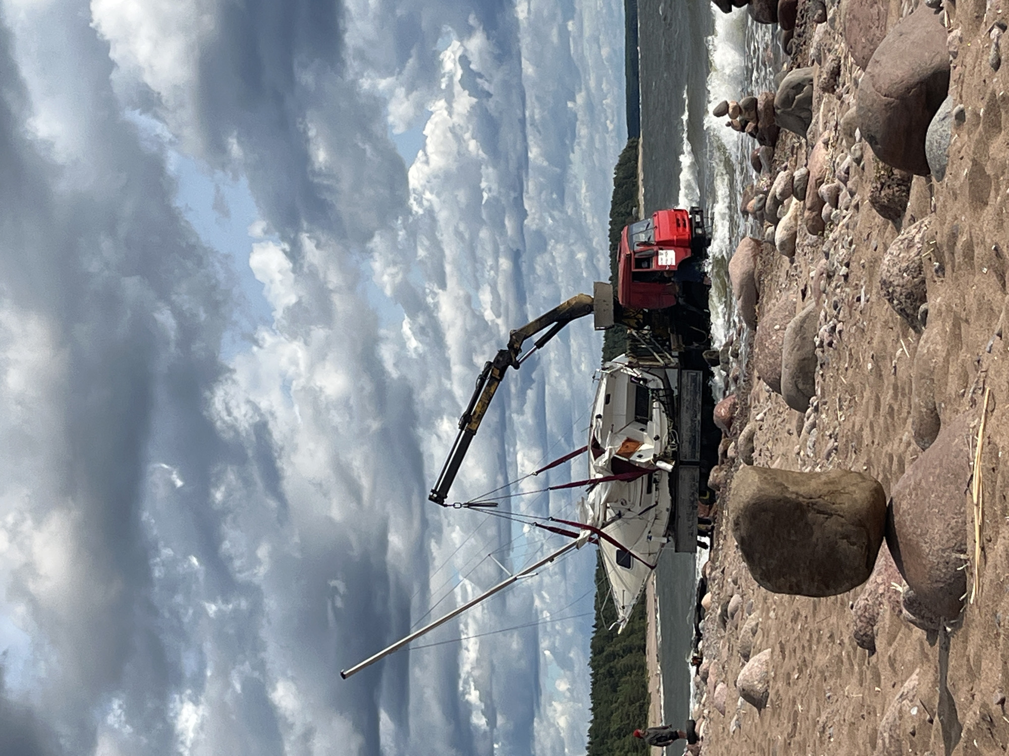 A sailboat is being lifted by a crane attached to a red truck on a sandy beach. The sailboat appears damaged and is being moved from the water onto land. The scene is set against a backdrop of a choppy sea, a treeline, and a cloudy sky. Several large rocks are scattered on the beach, and a few cairns are visible near the water. A person wearing a red hat stands on the left side of the frame, observing the operation. The overall impression is one of a salvage or recovery operation taking place in potentially inclement weather.