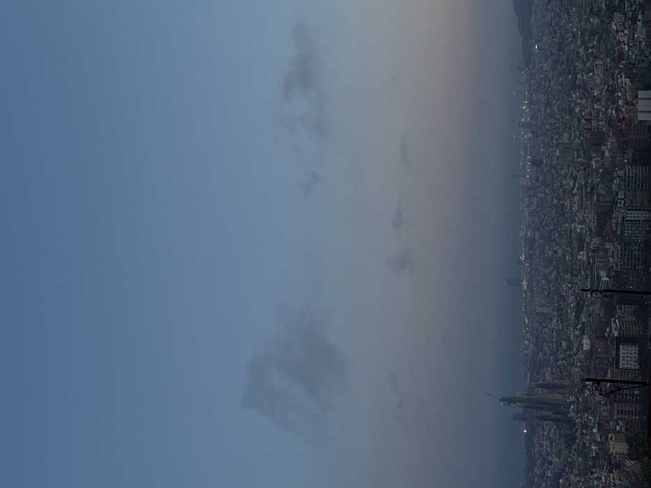 The image displays an elevated panoramic view of the city of Barcelona, Spain, observed at twilight. The upper two-thirds of the frame consist of a sky transitioning from a light blue at the top to a muted grey-purple closer to the horizon, with sparse, wispy grey clouds. The lower third reveals a sprawling, densely built urban landscape extending towards the Mediterranean Sea, which is visible as a dark band along the distant horizon. The Sagrada Familia basilica, characterized by its distinctive spires and a visible construction crane, is prominent in the lower-left quadrant of the cityscape. Numerous small, diffused points of artificial light are visible across the urban area and along the distant coastline, indicating the transitional lighting conditions. The immediate foreground contains two dark, indistinct vertical structures, possibly poles. No individuals or specific activities are discernible.