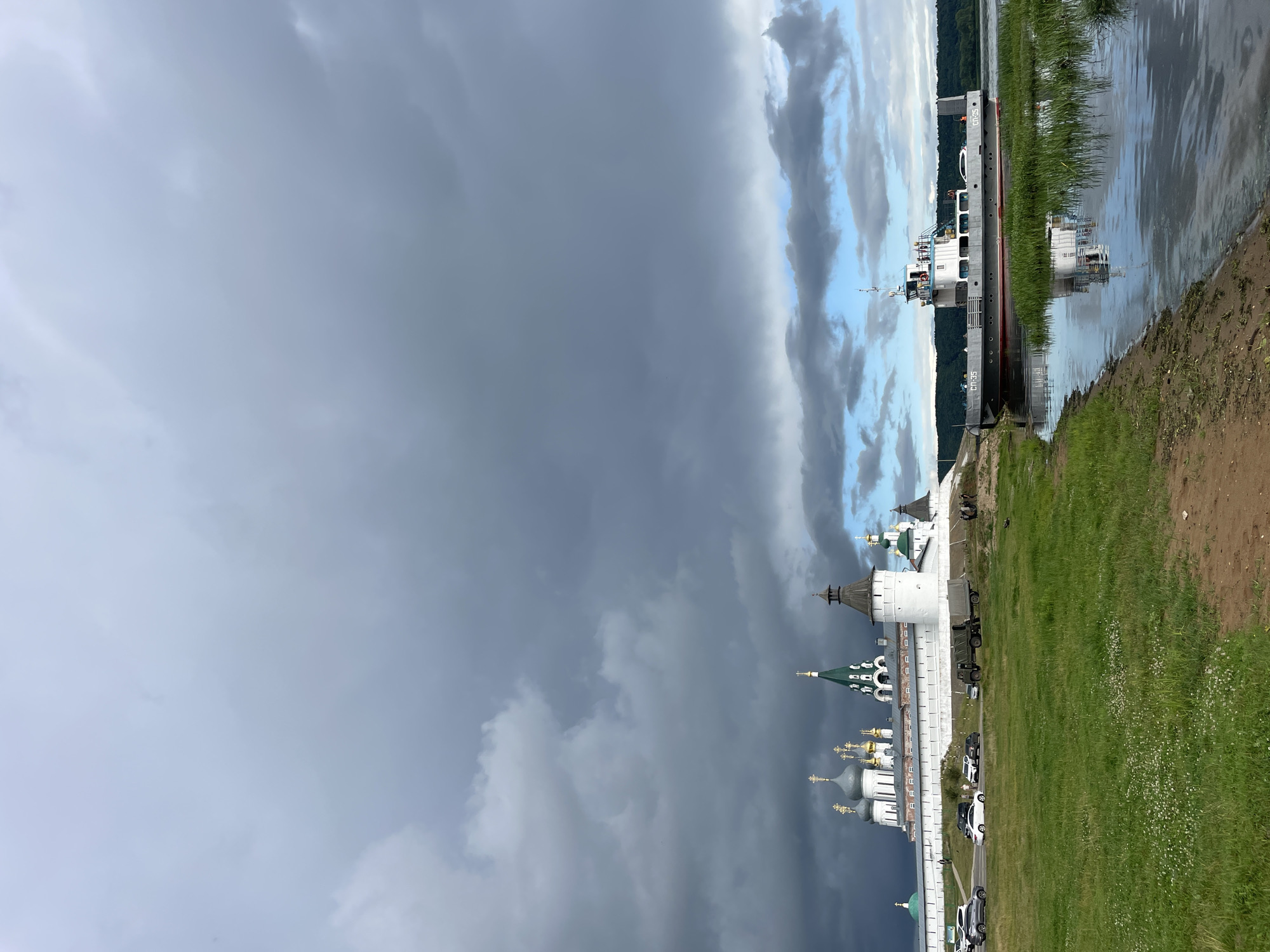 This image depicts the Trinity Monastery of Saint Macarius in Makar'evo, Russia, situated on the banks of a wide river. The monastery complex is characterized by its white stone walls, multiple domed churches with gold and green spires, and defensive towers. In the foreground, a ferryboat with the designation "СП-35" is docked near the grassy bank. The river reflects the boat and the sky, which is filled with dramatic, dark, and light grey clouds, suggesting an overcast or approaching storm. On the bank near the monastery, several vehicles are parked, including cars and a military-style truck. A few individuals are visible near the vehicles and along the monastery walls. The landscape includes green grass and a sandy shoreline.