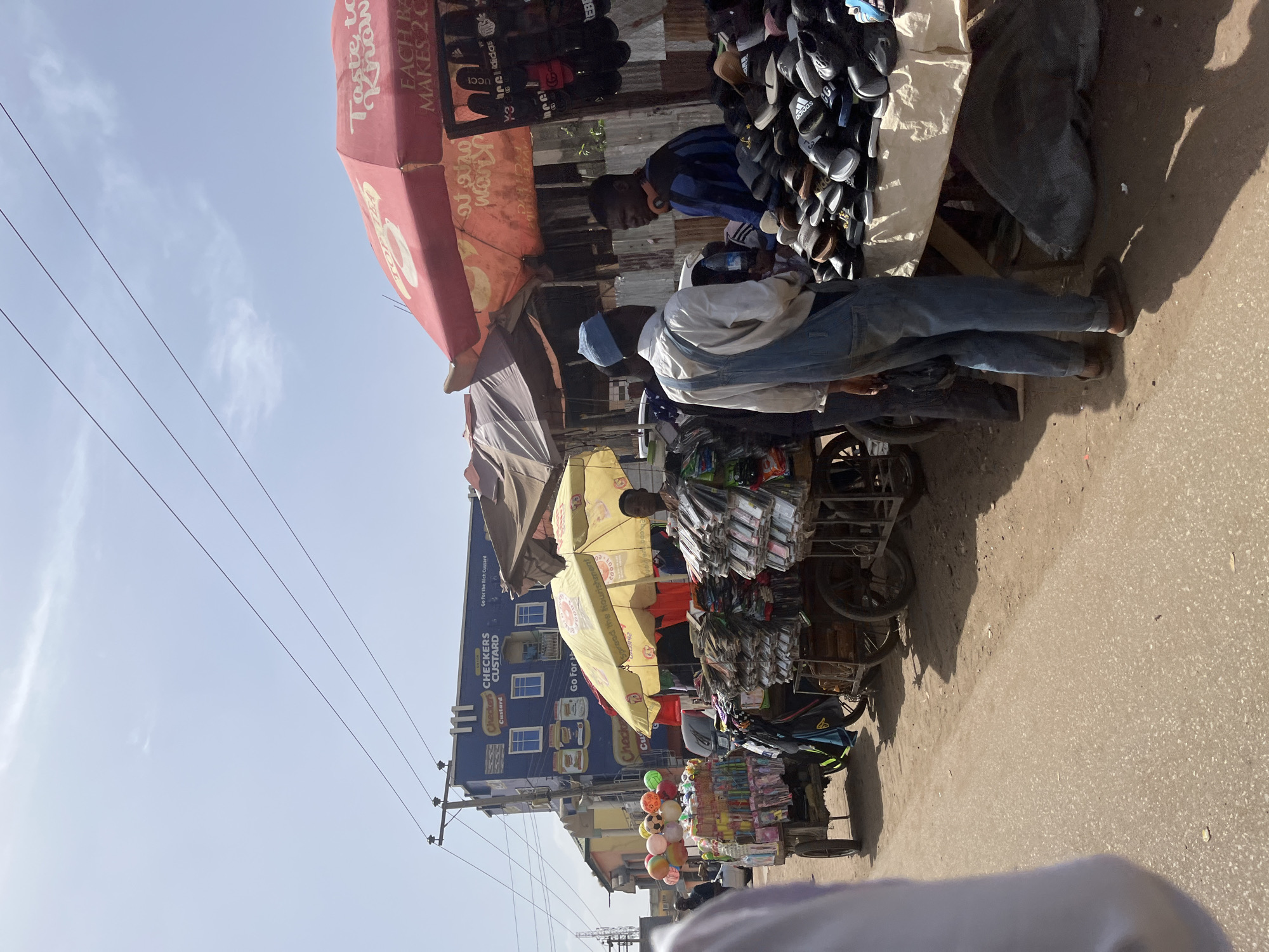 The image shows a street scene in Jos, Nigeria. A series of vendors are selling goods along the road. One vendor is selling shoes and sandals under a red and orange umbrella. Another vendor is selling cell phone accessories and phone cases from a cart under a yellow umbrella. There are also vendors selling balloons and other small goods. There are people browsing and interacting with the vendors. In the background, there's a building with a "Checkers Custard" sign. Power lines stretch across the top of the frame. The scene appears to be taken during the daytime with clear skies.