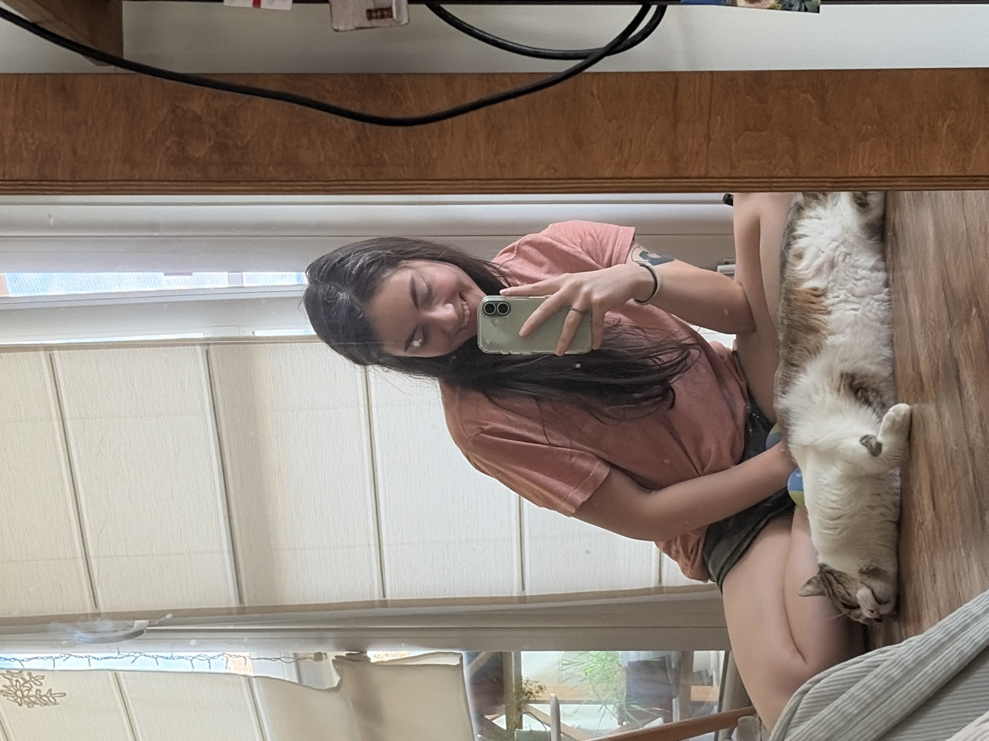 A young woman sits on the floor, taking a selfie in a mirror. She is wearing a pink t-shirt and dark shorts. A cat is lying next to her. The room is decorated with blinds and plants. The woman is smiling at the camera. The cat is asleep.