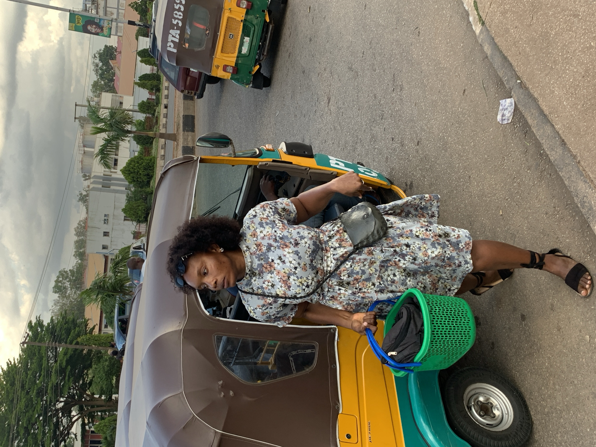 This image shows a woman standing next to a yellow and green auto-rickshaw (also known as a "keke"). She is wearing a floral print dress and black sandals. A green plastic basket containing a black bag is placed on the ground next to the auto-rickshaw.
The scene appears to be a street in Nigeria, with other vehicles and buildings visible in the background. The sky is cloudy, suggesting it might be overcast or early morning/late afternoon. The auto-rickshaw has the text "PTA-5859" and possibly "PETROL" visible on its exterior. The woman's expression is neutral, and she seems to be looking towards the front of the auto-rickshaw. The overall impression is of a street scene, possibly a moment of transit or waiting.
