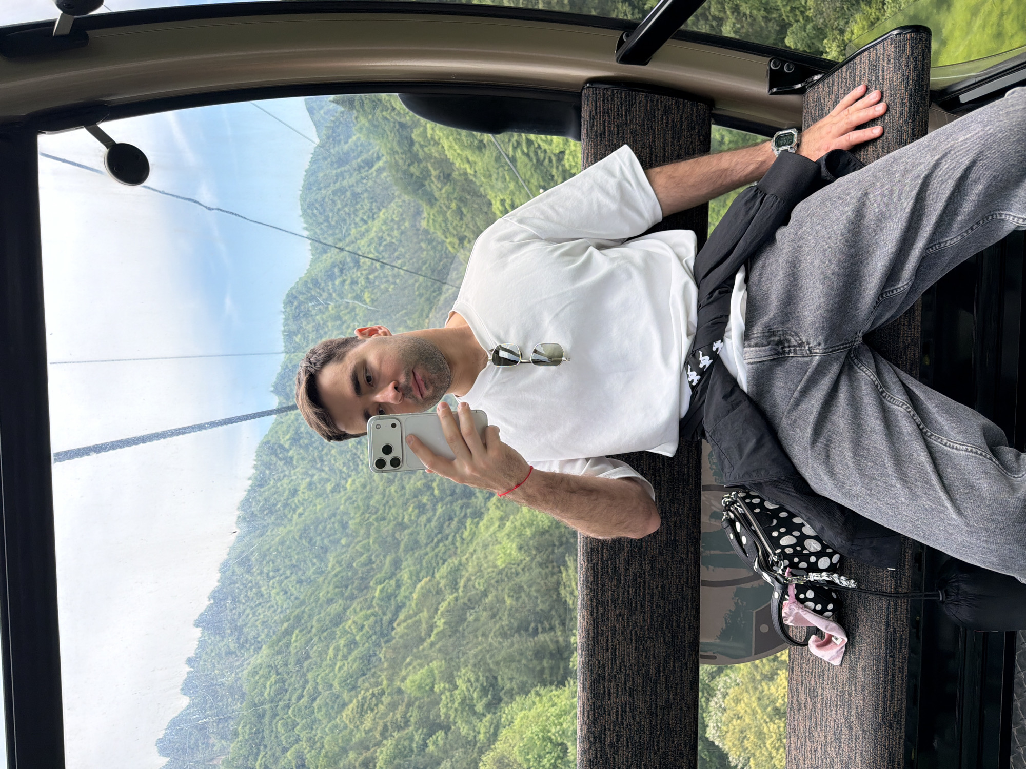 A man is inside a cable car, taking a selfie with his phone. The backdrop outside the cable car window shows the landscape of Wulingyuan, China, characterized by its distinctive sandstone pillars covered in lush greenery. The man is wearing a white t-shirt and gray jeans. Sunglasses are tucked into the collar of his shirt. He is holding a white smartphone to take the photo. A black jacket and a black and white patterned bag are situated beside him on the seat. The cable car interior consists of brown upholstered seating and large windows allowing for panoramic views.