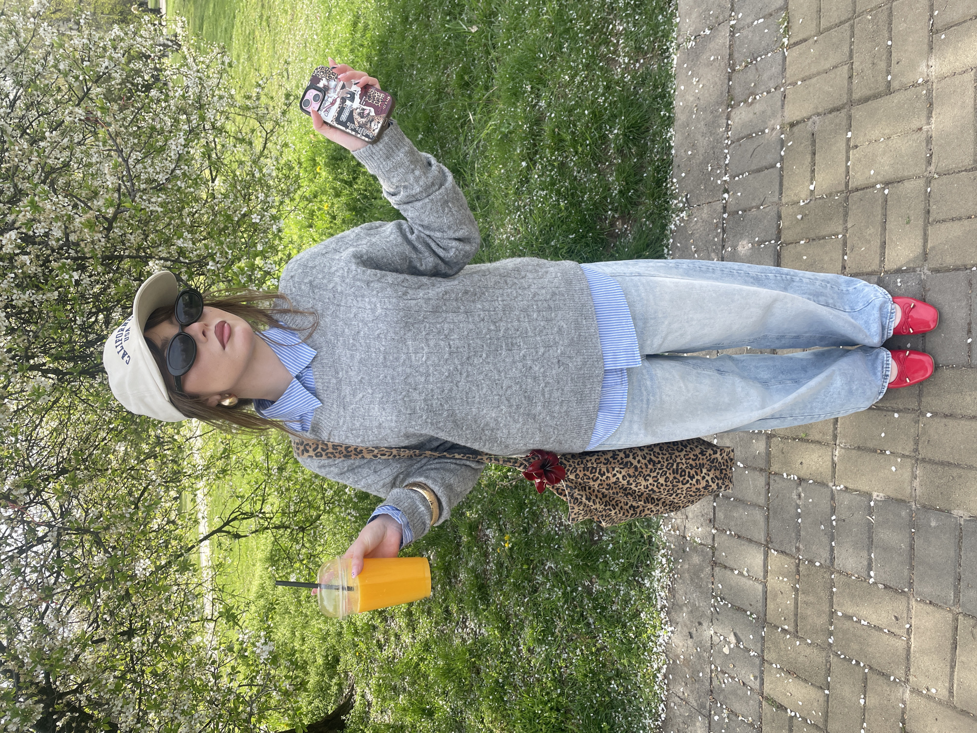 A full-body image, oriented vertically, depicts a person standing outdoors on a paved pathway. The setting is likely a park or garden in Sofia, Bulgaria, on a bright, sunny spring day. White petals, possibly cherry blossoms, are scattered across the grey rectangular paving stones and the adjacent green grass, signifying the season.
The subject, appearing female, is dressed in a light grey cable knit sweater layered over a blue and white striped collared shirt. She wears light-wash, relaxed-fit jeans and striking red patent leather ballet flats. A cream or light beige baseball cap, partially revealing the text "CALIFO..." (likely "CALIFORNIA") on its front, is worn over her head, paired with dark sunglasses. She accessorizes with gold-toned hoop earrings and several bracelets on her right wrist.
In her raised left hand, the person holds a smartphone with a distinctive leopard print case adorned with various stickers, some cartoonish, and partial text like "VoD-vRo-allo" is visible. Her right hand holds a clear plastic cup containing an orange beverage, possibly juice or a smoothie, with a black straw. A leopard print shoulder bag, featuring a dark red fabric flower detail, hangs by her side. The background showcases vibrant green grass and trees in full bloom with white flowers. The overall scene conveys a casual and relaxed outing.