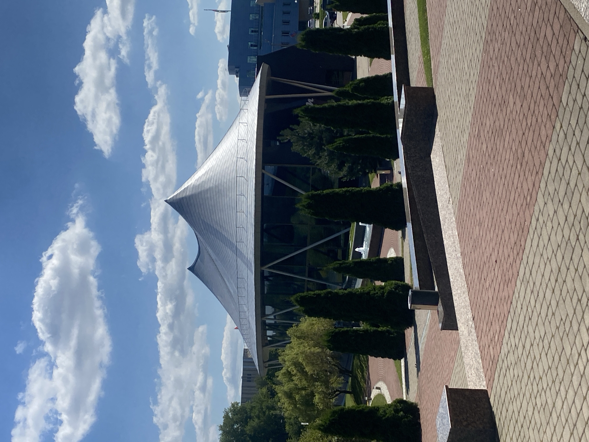 The image shows the National Library of Belarus in Minsk. The modern building has a distinctive geometric shape with a large, faceted facade. The library is surrounded by a landscaped area with trees, pathways, and a fountain. The sky is blue with scattered clouds.