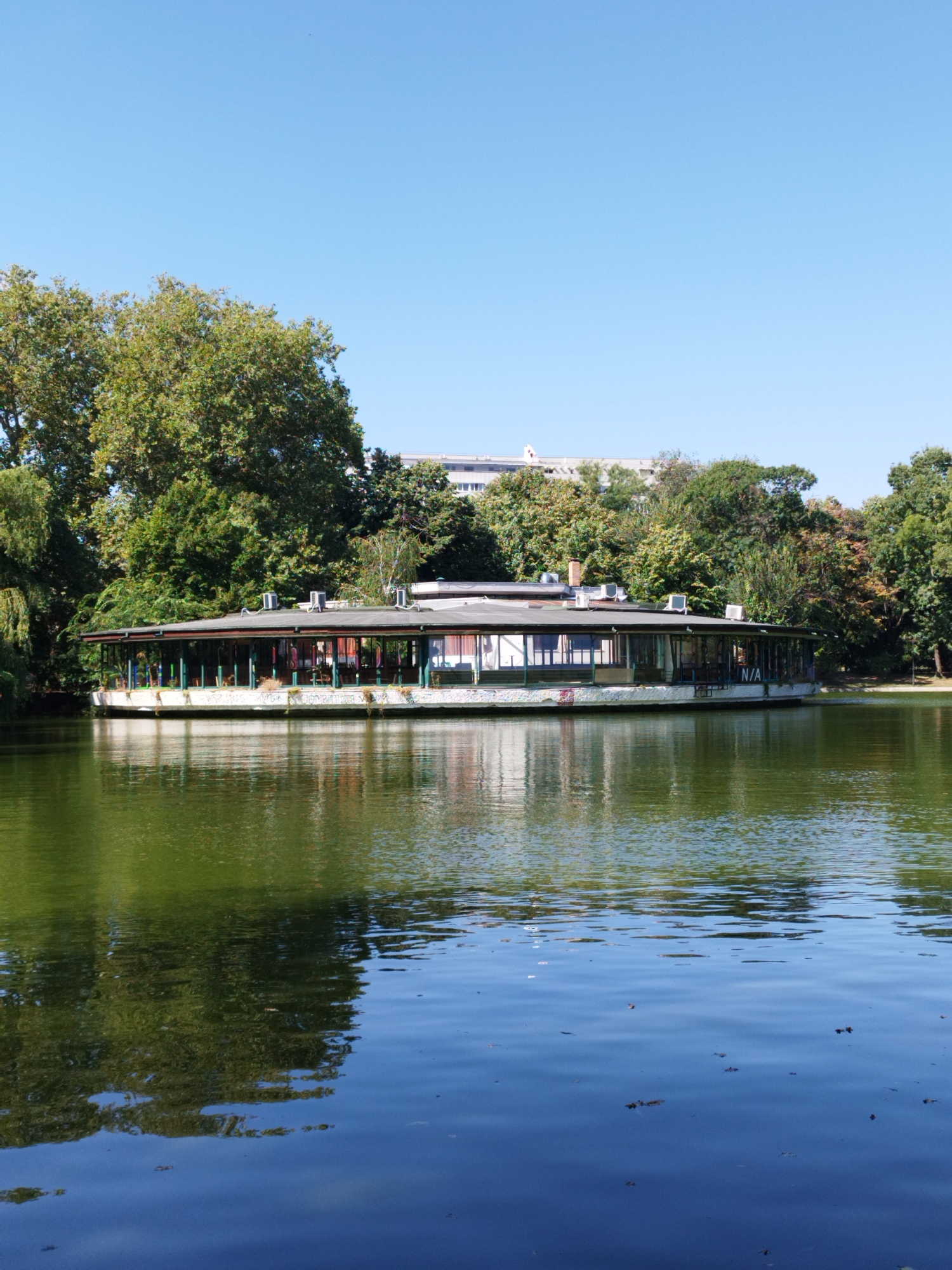 This is a vertical, full-shot, eye-level photograph of a building on a lake in Herăstrău Park (now King Michael I Park) in Bucharest, Romania. The building is a circular structure with a dark roof, an overhang, and glass walls. It appears to be a restaurant or cafe, with tables and chairs visible inside. The building is surrounded by trees, and the lake reflects the sky and trees. The sky is clear and blue, and the water is a greenish-blue color. The overall mood of the photograph is peaceful and serene.