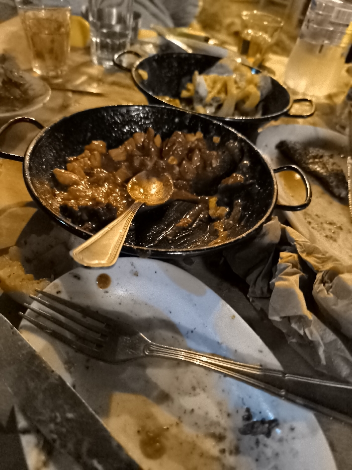 This media file captures a close-up, warm-toned view of a dining table in what appears to be a restaurant or tavern in Nea Erithrea, Greece, likely after a meal. The scene is dominated by remnants of food and used dishes.
In the foreground, a dark, round, shallow pan holds the last servings of a rich, brown, saucy dish with chunky pieces, possibly a stew or a hearty casserole. A golden, ornate spoon rests inside the pan. Below it, a dirty, light-colored plate is visible, upon which a silver fork with a decorative handle is placed, along with the edge of a knife.
Further back, another similar dark pan contains remnants of a lighter, yellowish dish, possibly fried potatoes or a pasta. Several glasses are scattered around the table; one holds a golden liquid (perhaps a drink), another is empty, and a taller glass or pitcher appears to contain water. A small, bright yellow piece of lemon is also discernible. Crumpled, stained paper napkins are strewn across the table surface.
No people are explicitly visible, though a blurred arm or shoulder is faintly seen in the far background, indicating diners were present. The warm, somewhat dim lighting suggests an evening setting, contributing to a casual and relaxed atmosphere. The array of serving pans and individual plates points to a shared meal. There is no visible text in the image.