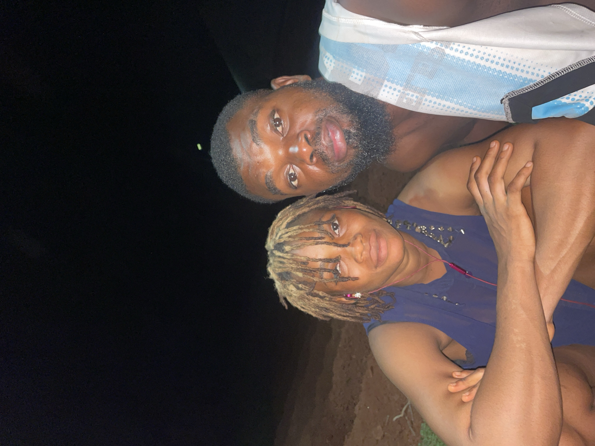Two people pose in an outdoor nighttime scene. The man on top has dark hair, a beard, and wears a light blue and white patterned shirt. Below him, a person with blonde dreadlocks wears a blue shirt and pink earbuds. The location appears to be in Ogbe, Nigeria, as indicated by the provided context. The background is dark, possibly a field or park.