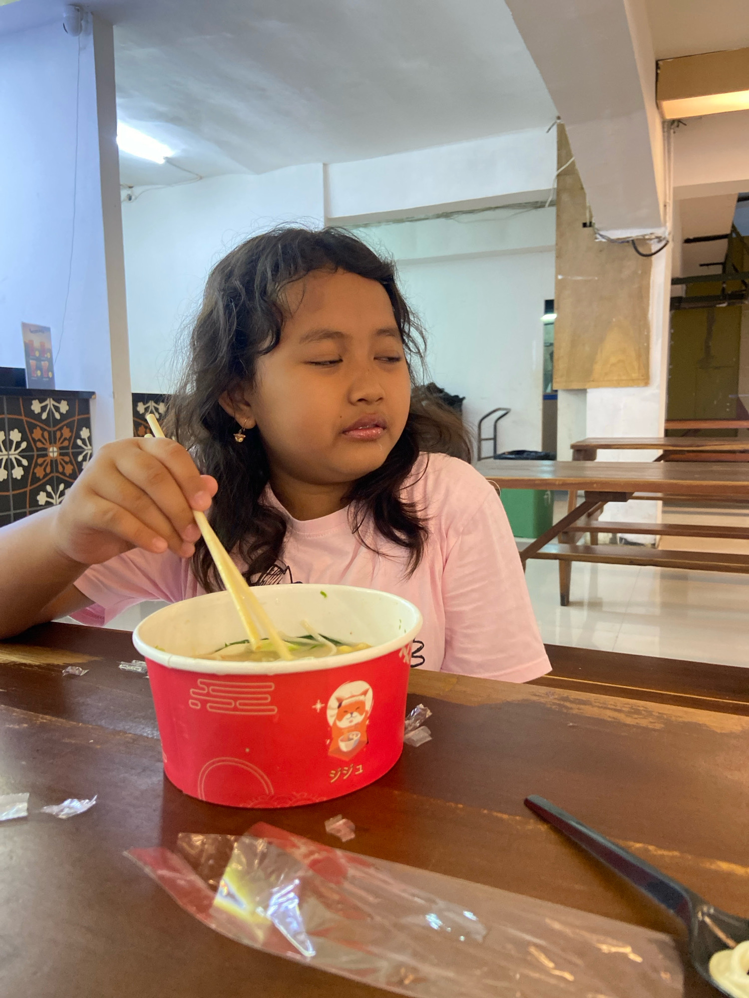 A young girl with curly dark hair is seated at a wooden table, holding chopsticks to eat from a red bowl of noodles. She is wearing a pink t-shirt. The background suggests an indoor dining area with tables and chairs. The setting appears to be a restaurant in Kecamatan Pati, Indonesia, based on the provided context. The girl's eyes are partially closed, and her mouth is slightly open as she eats. The table surface is made of dark wood and has some discarded plastic wrappers.
