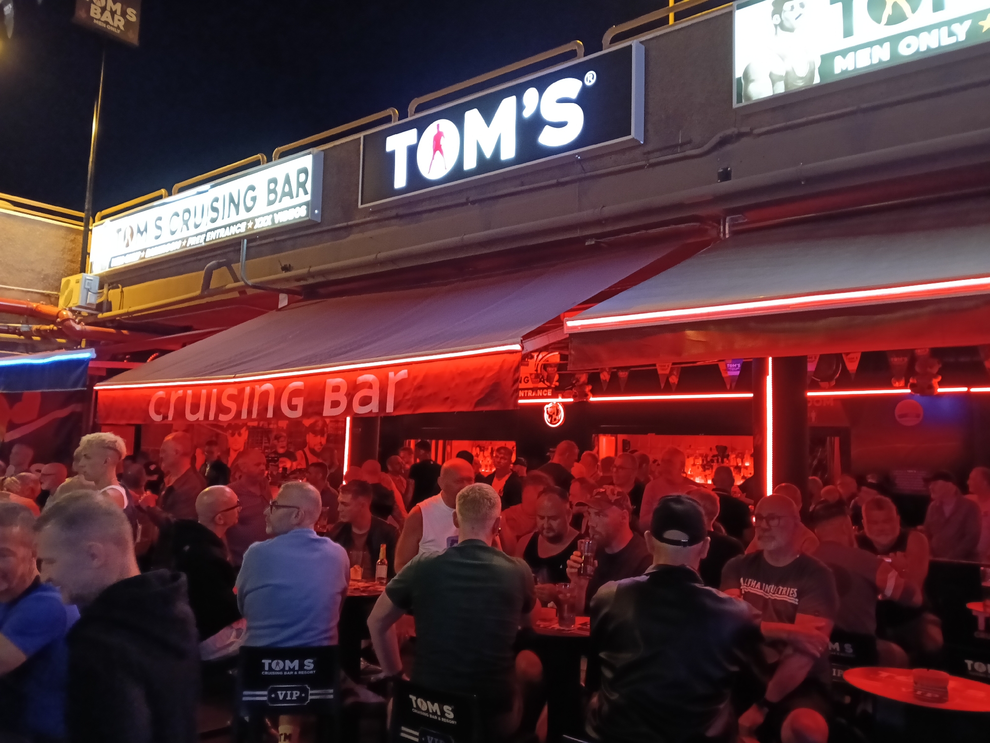 The image captures a lively nighttime scene outside and inside "Tom's Cruising Bar" in San Bartolomé de Tirajana, Spain. The bar is brightly lit, primarily with red neon lights that cast a warm, energetic glow on the patrons and interior. The sky above is dark, indicating it's evening or night.
The primary subjects are numerous men of varying ages, mostly in casual attire, socializing and drinking. Many are standing, while others are seated at tables or bars. The atmosphere appears to be busy and social, with people engaged in conversations. The red lighting permeates the interior, highlighting the faces and figures of the men, some of whom are holding drinks.
Notable details include prominent signage. Above the awning, a large illuminated sign reads "TOM'S" with a stylized figure of a man within the "O." Another sign clearly states "TOM'S CRUISING BAR," with smaller text below indicating "MEN ONLY," "FREE ENTRANCE," and "XXX VIDEOS." A smaller sign further up reiterates "TOM'S BAR MEN ONLY." On the chairs, text includes "TOM S CRUISING BAR & RESORT" and "VIP." One man is seen wearing a t-shirt with "ALPHA INDUSTRIES" visible. The bar features a retractable awning or roof, underneath which the "cruising Bar" text is visible, also in red neon. The space appears to be a combination of an outdoor patio area and an indoor section.