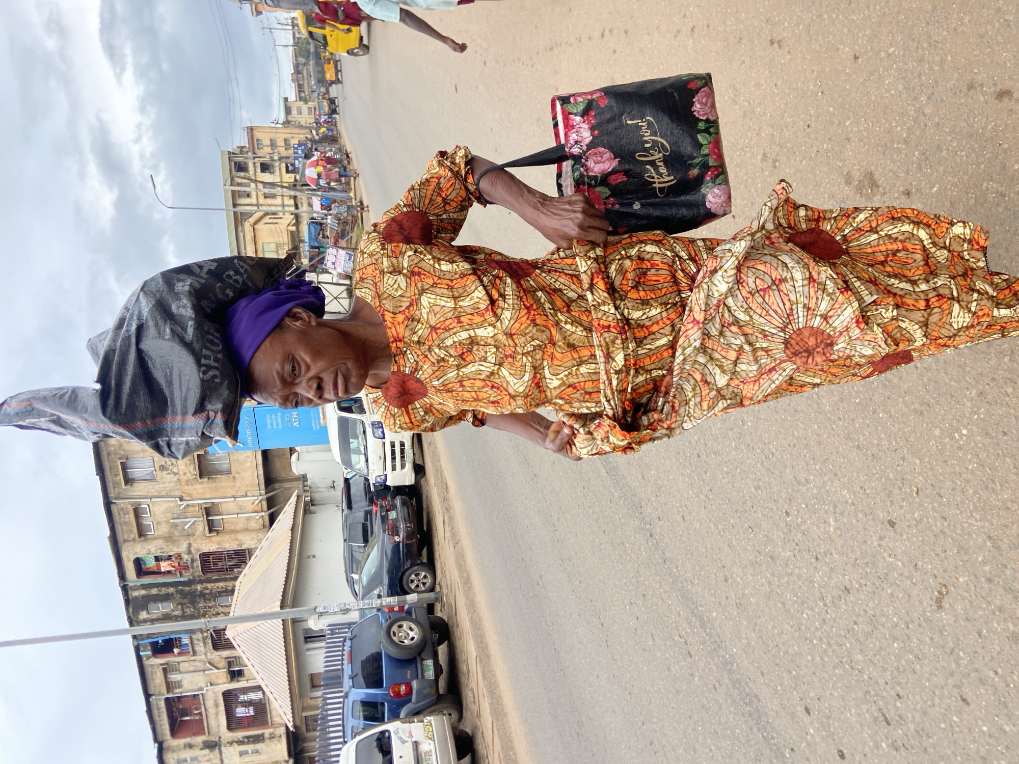 In Nkpor, Nigeria, an elderly woman walks down a paved road with a large black plastic bag balanced on her head. She is wearing a vibrant orange and brown patterned dress and a purple headwrap. In her right hand, she carries a black tote bag with a floral design and the words "Thank you!" printed in gold script. Several vehicles, including a blue Jeep and a white van, are parked along the side of the road. In the background, there are multi-story buildings, suggesting an urban or semi-urban environment. The sky is overcast, indicating a cloudy day.