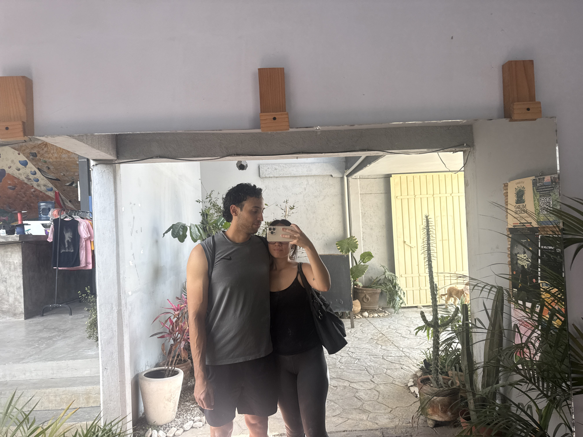 A man and a woman are standing together in front of a large mirror, taking a selfie. The man is wearing a gray tank top and dark shorts. The woman is wearing a black tank top and dark leggings, holding her smartphone to take the photo. The mirror reflects an outdoor area with plants in pots, a gate in the background, and glimpses of the surroundings. Based on the decor and style of the building reflected in the mirror, the location is likely a cafe or shop in San Andrés Cholula, Mexico.