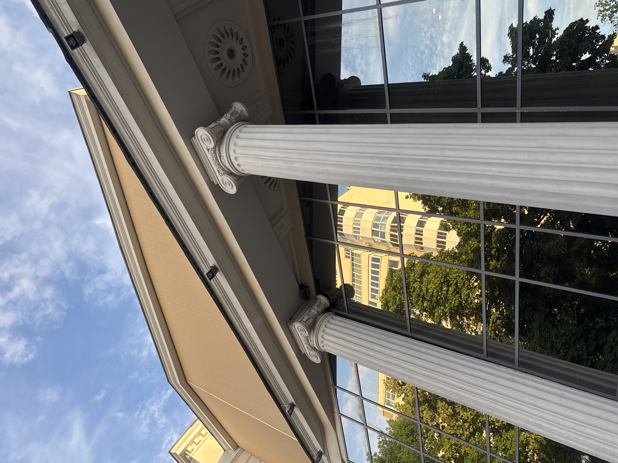 The image shows a close-up, low-angle view of the facade of a building with classical architectural elements, located in Novaya Usman', Russia. The building features large, reflective glass windows and white, fluted columns with ornate Corinthian capitals. Above the columns, the building's exterior is painted a light tan color. The glass windows reflect the surrounding environment, including a multi-story building with many windows, green trees, and the blue sky with scattered white clouds. The reflection suggests an urban setting. The overall impression is one of a modern building integrated with classical design, possibly a public or commercial establishment given its grand facade.