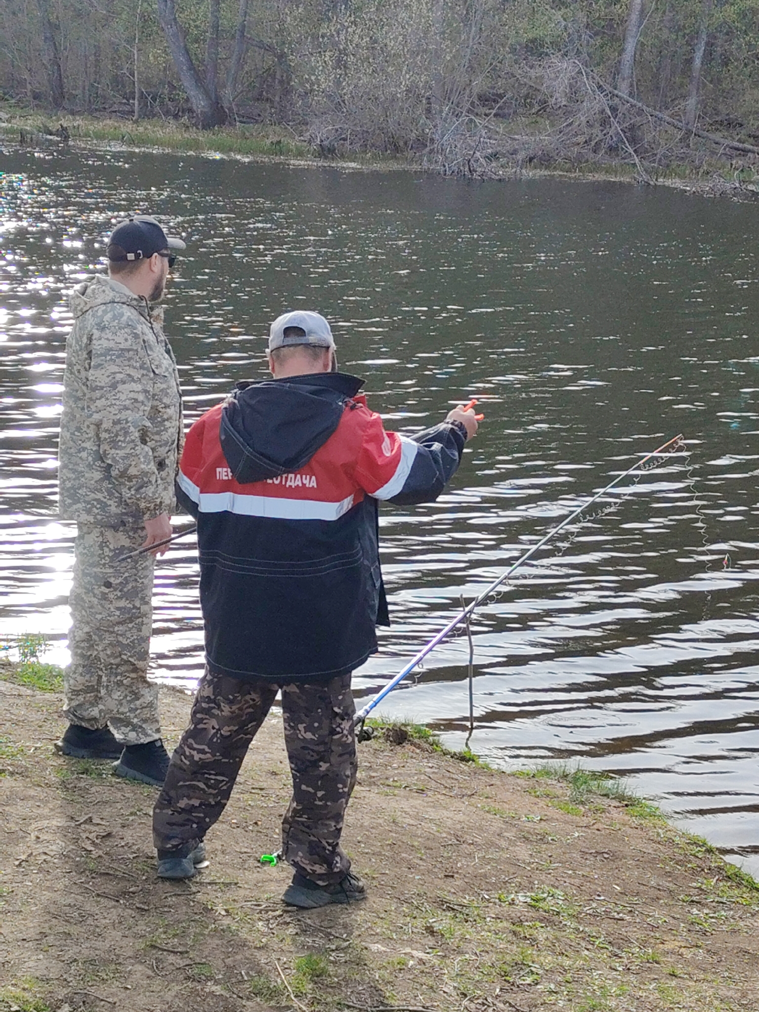 Two men are fishing at the edge of a body of water. One man is wearing camouflage clothing, while the other is wearing a red, black, and white jacket with camouflage pants. The man in the jacket is pointing towards the water, while holding a fishing rod. A second fishing rod is propped against the bank. The water reflects the sunlight, and trees line the opposite shore. The location is Unknown, Unknown.