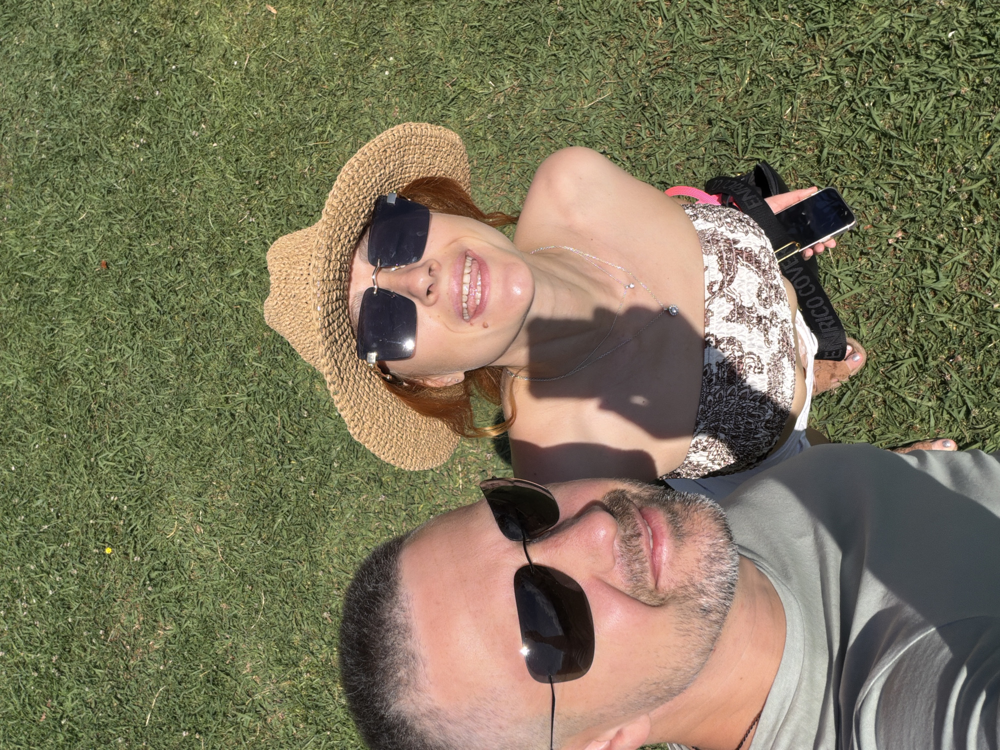 An overhead photograph captures two individuals, a man and a woman, lying on a vibrant green grass lawn under bright sunshine in Budva, Montenegro. The image is taken from a high angle, with the subjects oriented diagonally across the frame; their heads are positioned towards the lower right, and their bodies extend upwards to the left.
The woman, located in the upper right portion of the image, is smiling broadly at the camera. She wears a straw hat and dark, rectangular sunglasses. Her reddish-brown hair is visible beneath the hat. She has a delicate silver necklace with a small pendant around her neck and is dressed in a light-colored top with a brown and white patterned design. Her left hand holds a black smartphone, and a black bag strap with white text, partially reading "RICKO COVE" and "ENBE," is visible across her torso.
The man is positioned in the lower left, partially overlapping the woman. He has short dark hair, a beard, and is wearing dark rectangular sunglasses. He looks towards the camera with a relaxed expression. He is dressed in a light green t-shirt and a dark beaded necklace.
The strong sunlight creates distinct shadows across their bodies and faces, suggesting a clear, sunny day, likely mid-day or afternoon. The green grass indicates an outdoor, leisure setting. The woman's wide smile conveys a sense of happiness and enjoyment.