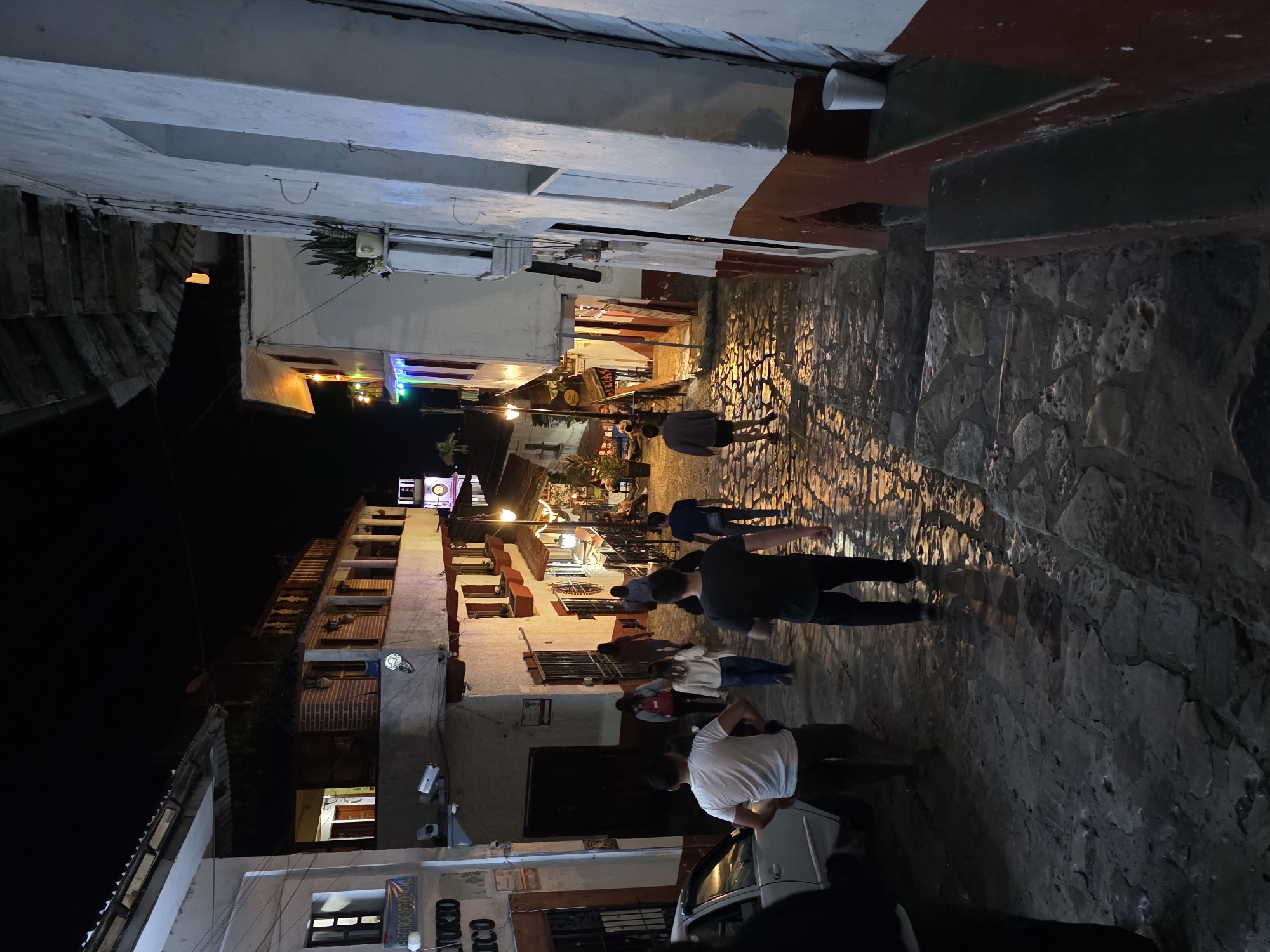 The image shows a narrow, cobblestone street at night in Cuetzalan, Mexico. The street is lined with two and three-story buildings that are predominantly white with red trim. Several people are walking along the street, their figures illuminated by streetlights and lights emanating from the buildings. The street appears to be sloping upwards from the bottom of the frame. A car is parked on the right side of the street at the bottom of the frame. The sky is dark and visible above the buildings.